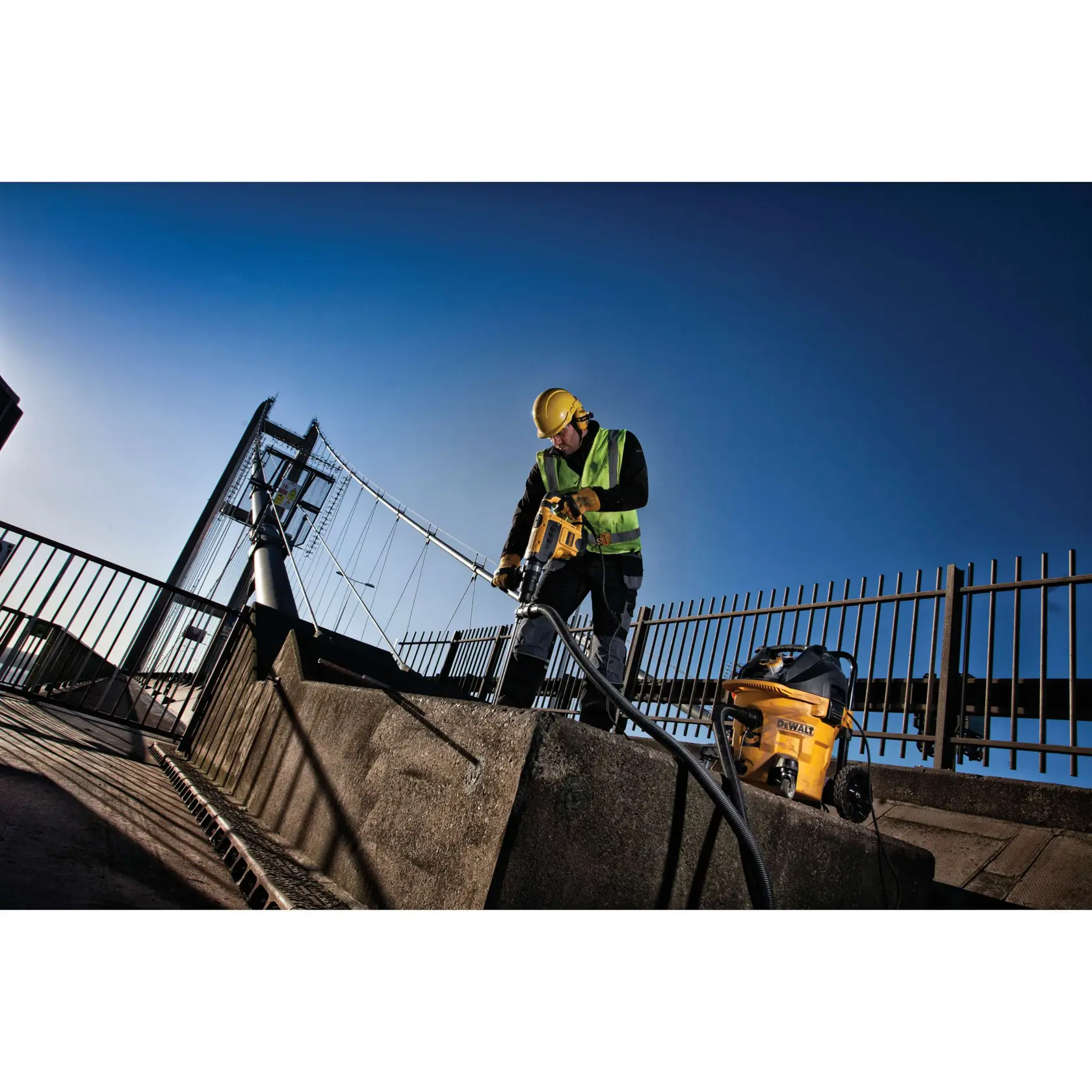 Trabajador usa herramienta DEWALT en concreto junto al puente, lleva casco y tiene equipo DEWALT cerca.