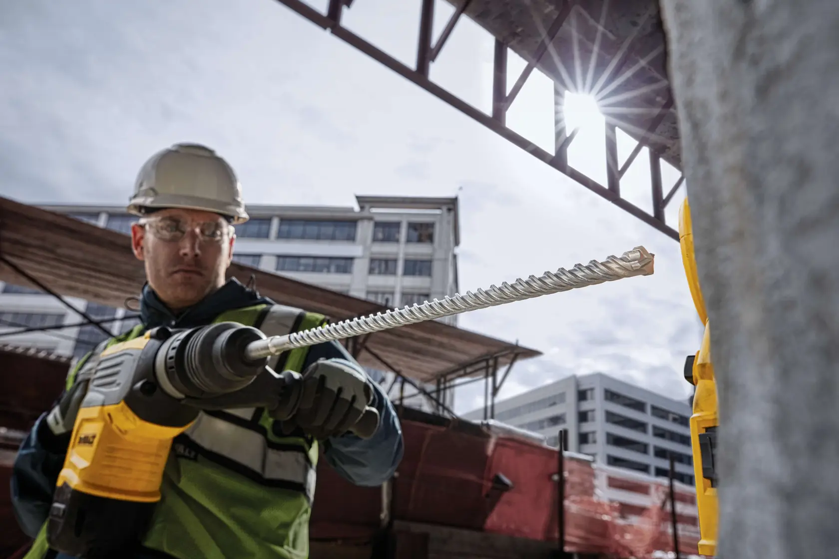 Trabajador usa taladro DEWALT con broca larga en obra, edificios de fondo.