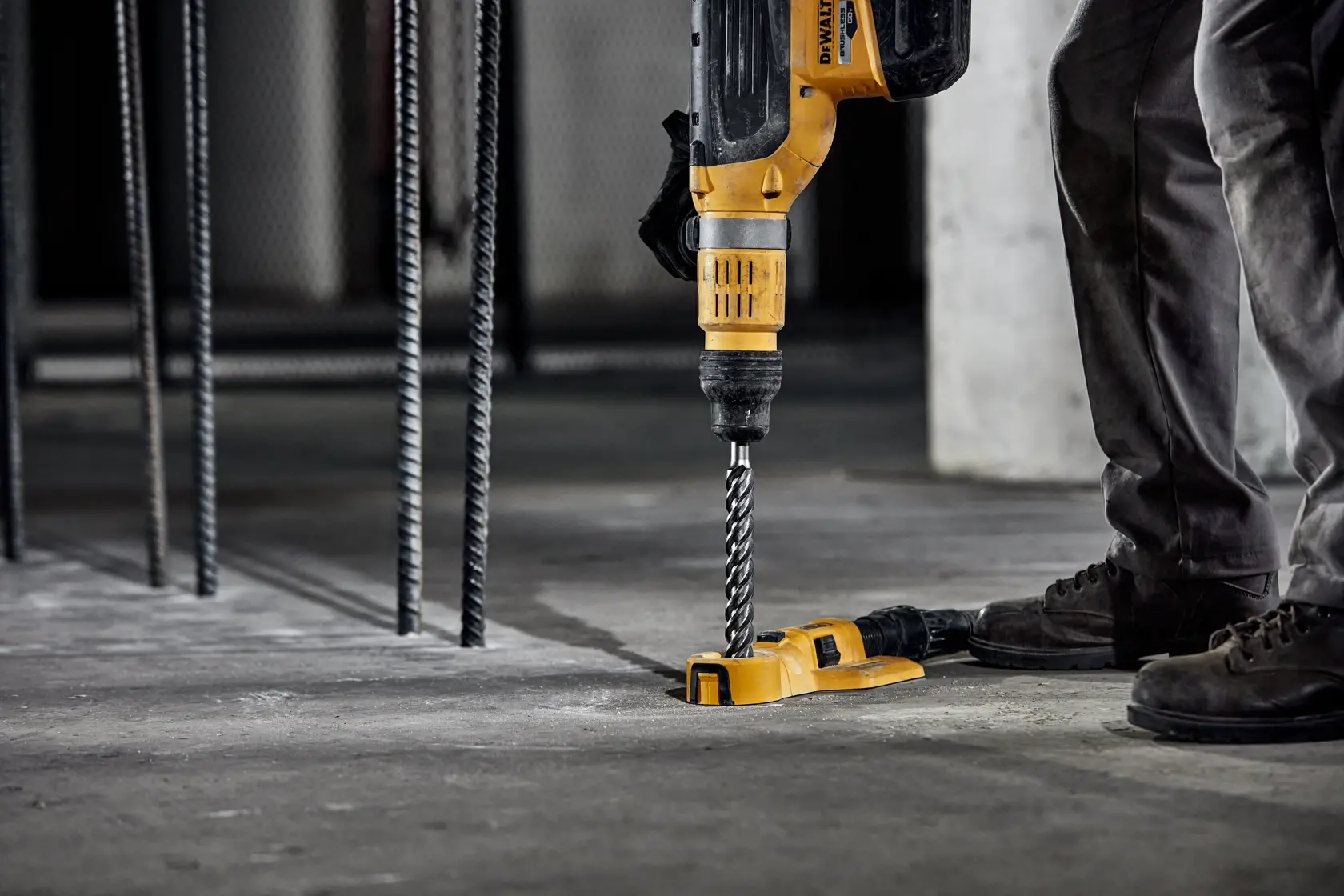 Person nutzt DEWALT Schlagbohrmaschine mit Bohrer auf Betonboden an Baustelle.