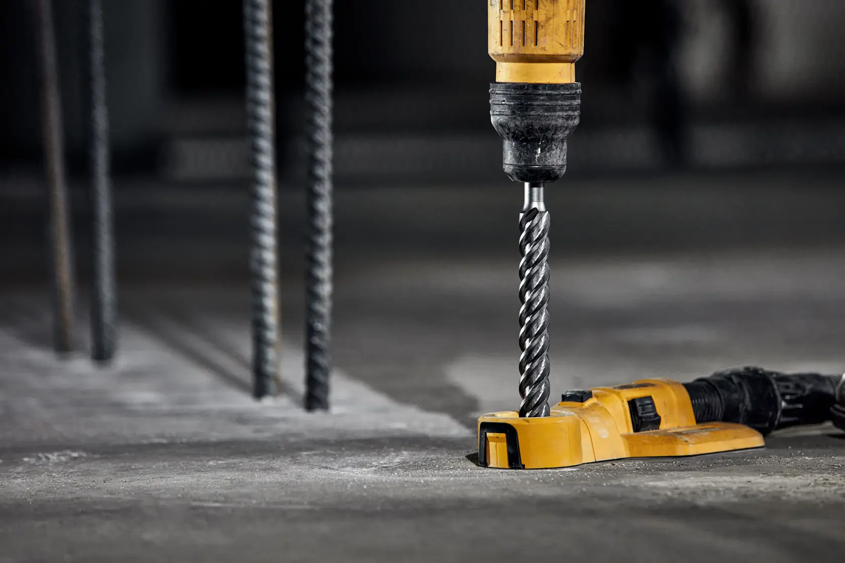 Ein gelber DEWALT-Bohrer mit einem Spiralbohrer wird verwendet, um in eine Betonoberfläche zu bohren. Metallstangen sind im Hintergrund zu sehen, was auf eine Baustellenumgebung hindeutet.