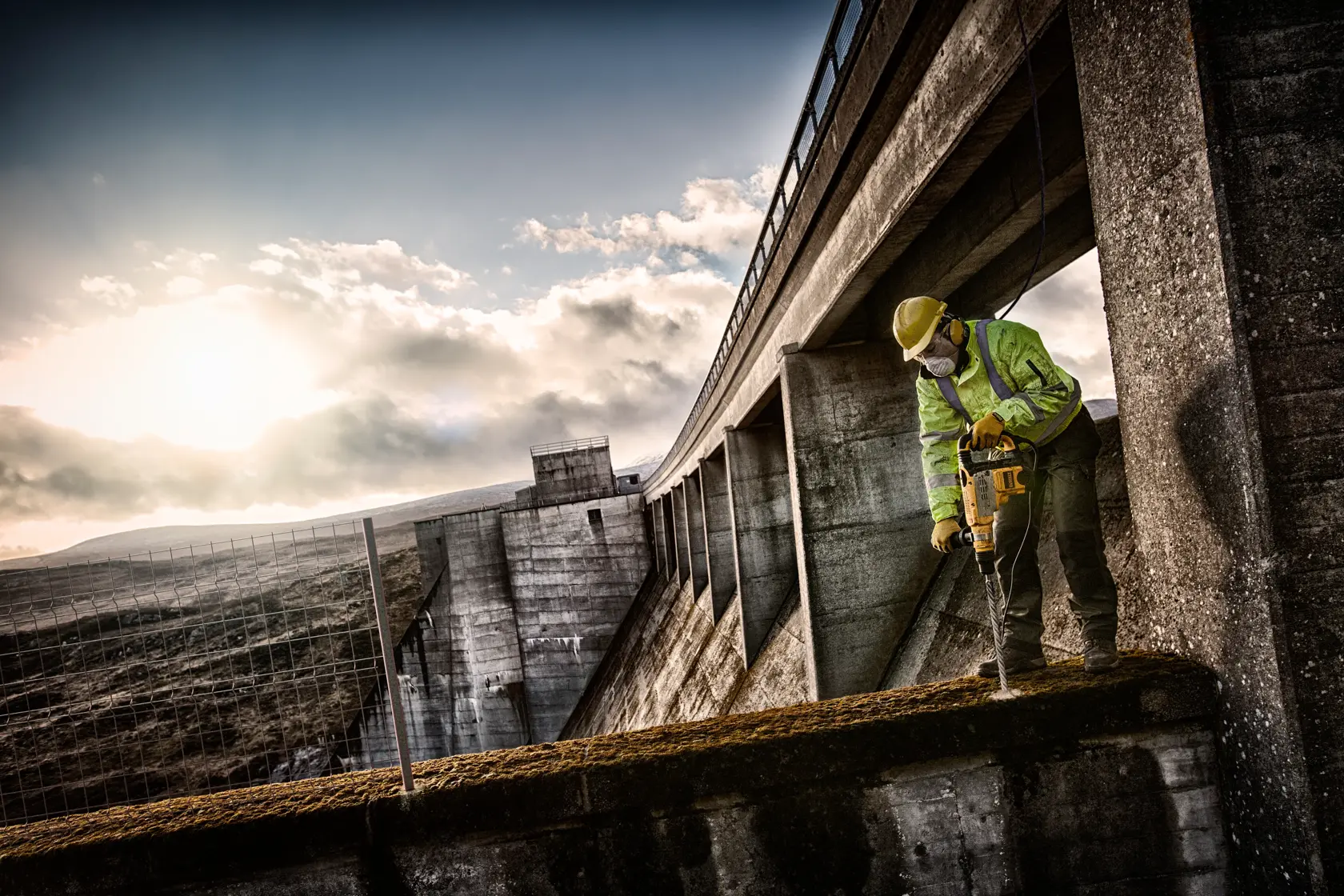 Arbeiter nutzt Dewalt-Bohrmaschine an Beton bei Sonnenaufgang, mit Schutzkleidung und Helm.