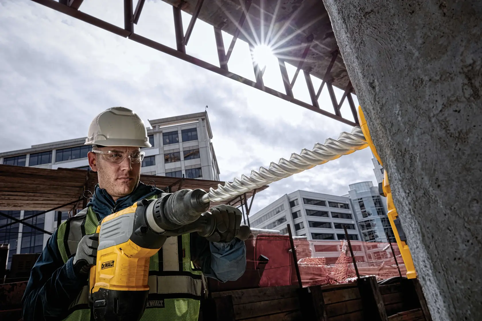 Trabajador usa taladro DEWALT con broca espiral en concreto en obra, edificios modernos al fondo.