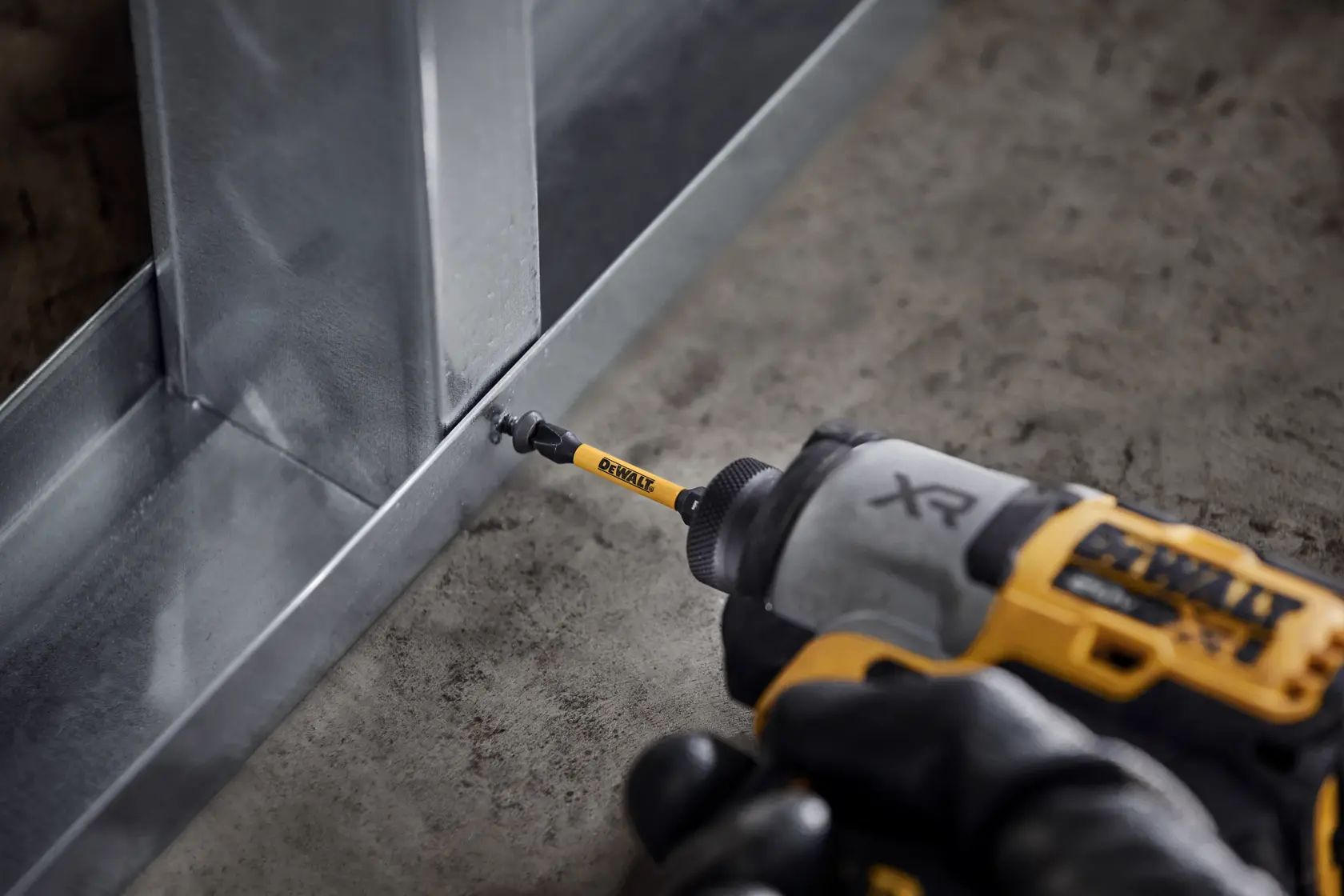 DEWALT XR drill driving screw into metal frame on a concrete floor, close-up of tool and bit in action.