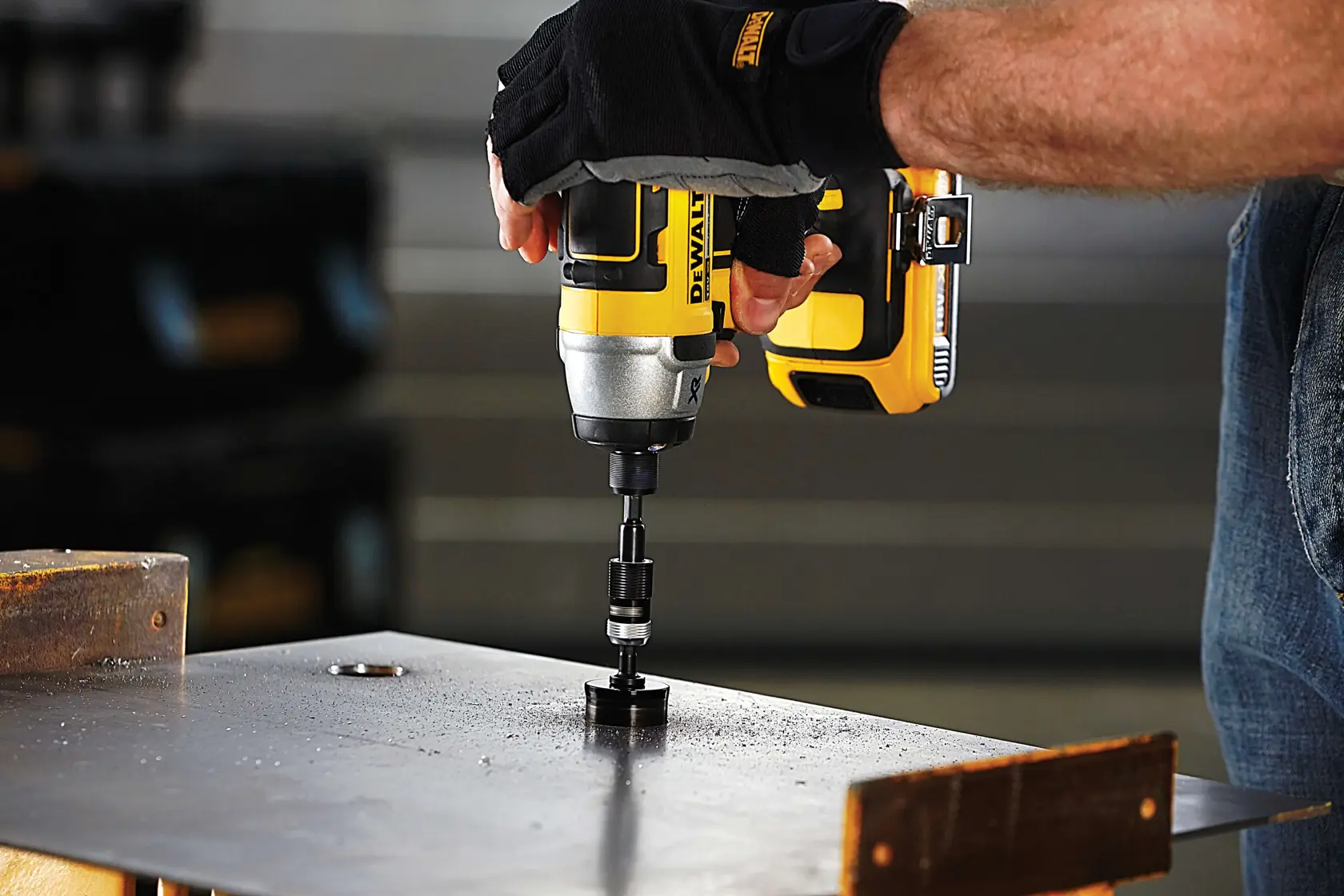 Gloved hand uses a DEWALT drill with hole saw attachment on a metal sheet in a workshop setting.