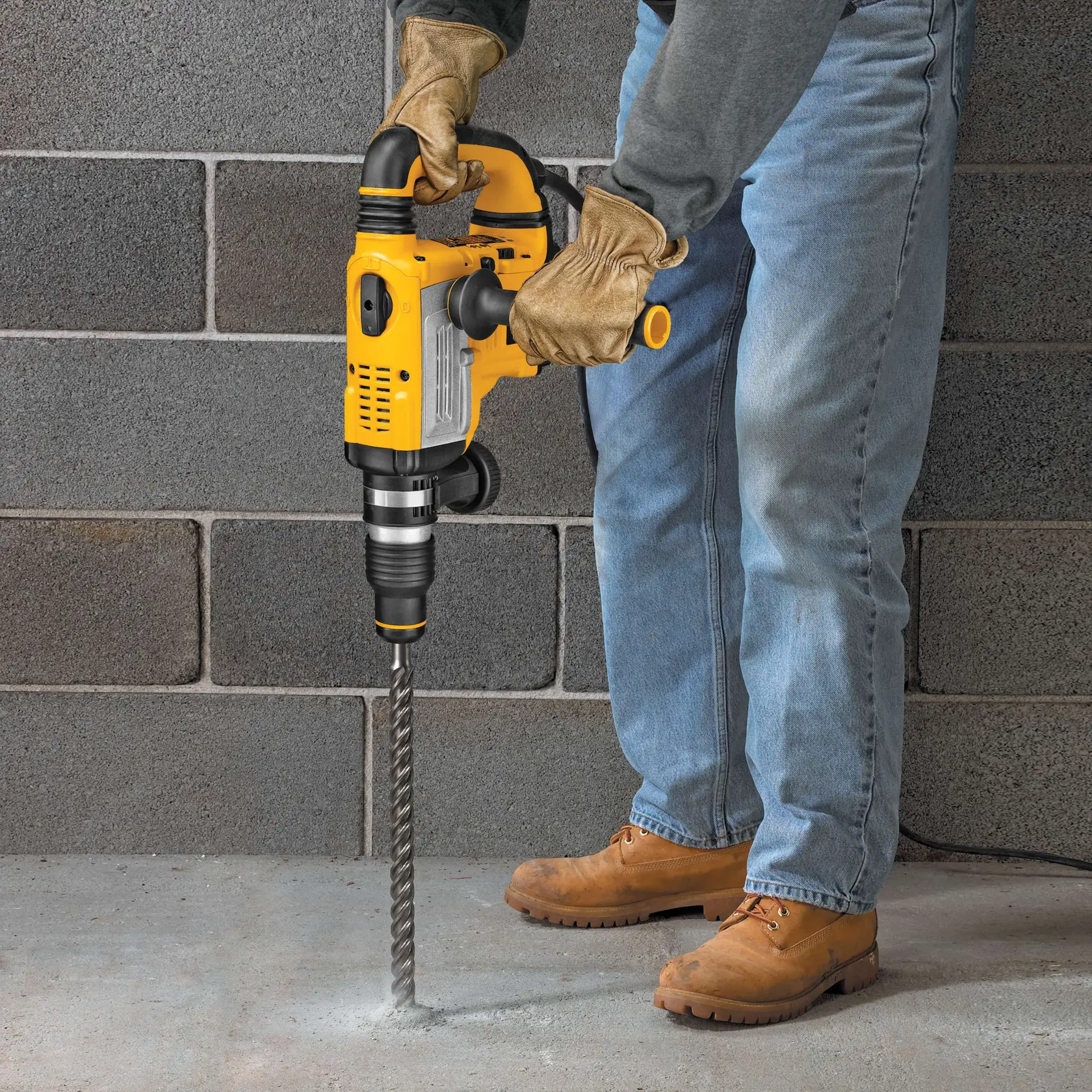 Person bohrt Betonboden mit gelbem DEWALT-Bohrhammer, trägt Handschuhe und Stiefel.