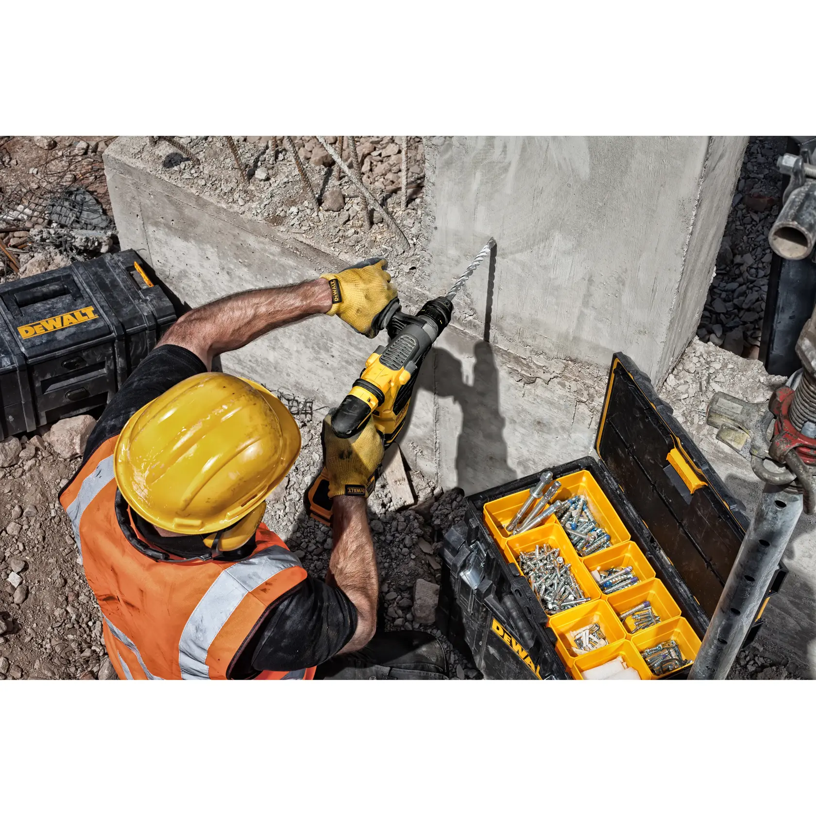 Trabajador perfora pared de cemento con herramienta DEWALT junto a caja abierta de fijaciones.