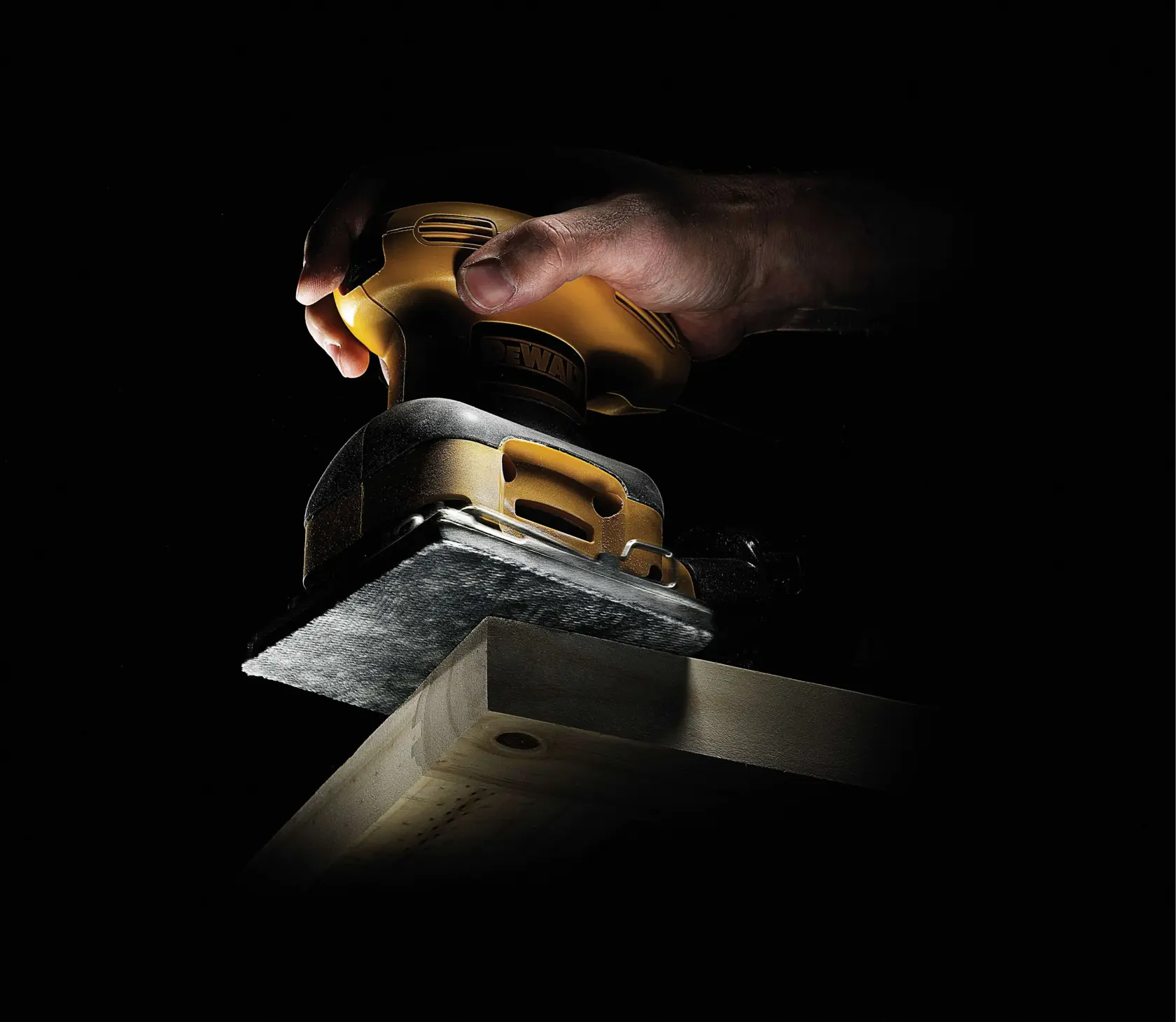 Mano usando una lijadora DEWALT en una tabla de madera con poca luz.