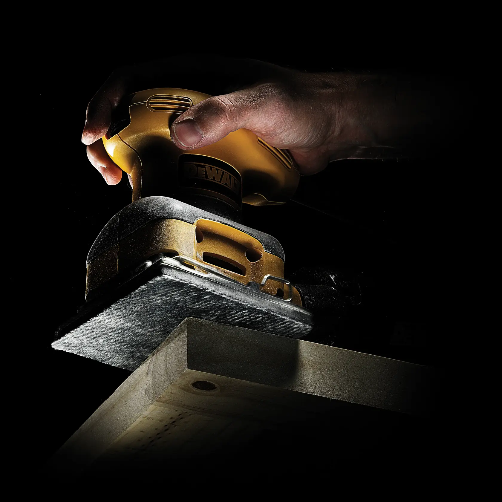 Mano usando lijadora DEWALT, suaviza una superficie de madera bajo luz focalizada.