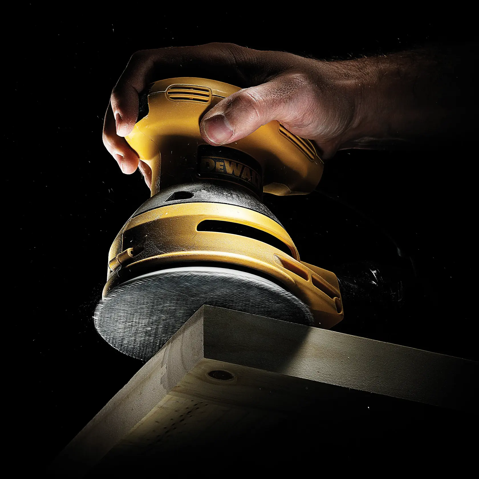Mano sostiene lijadora DEWALT puliendo borde de madera sobre fondo oscuro.