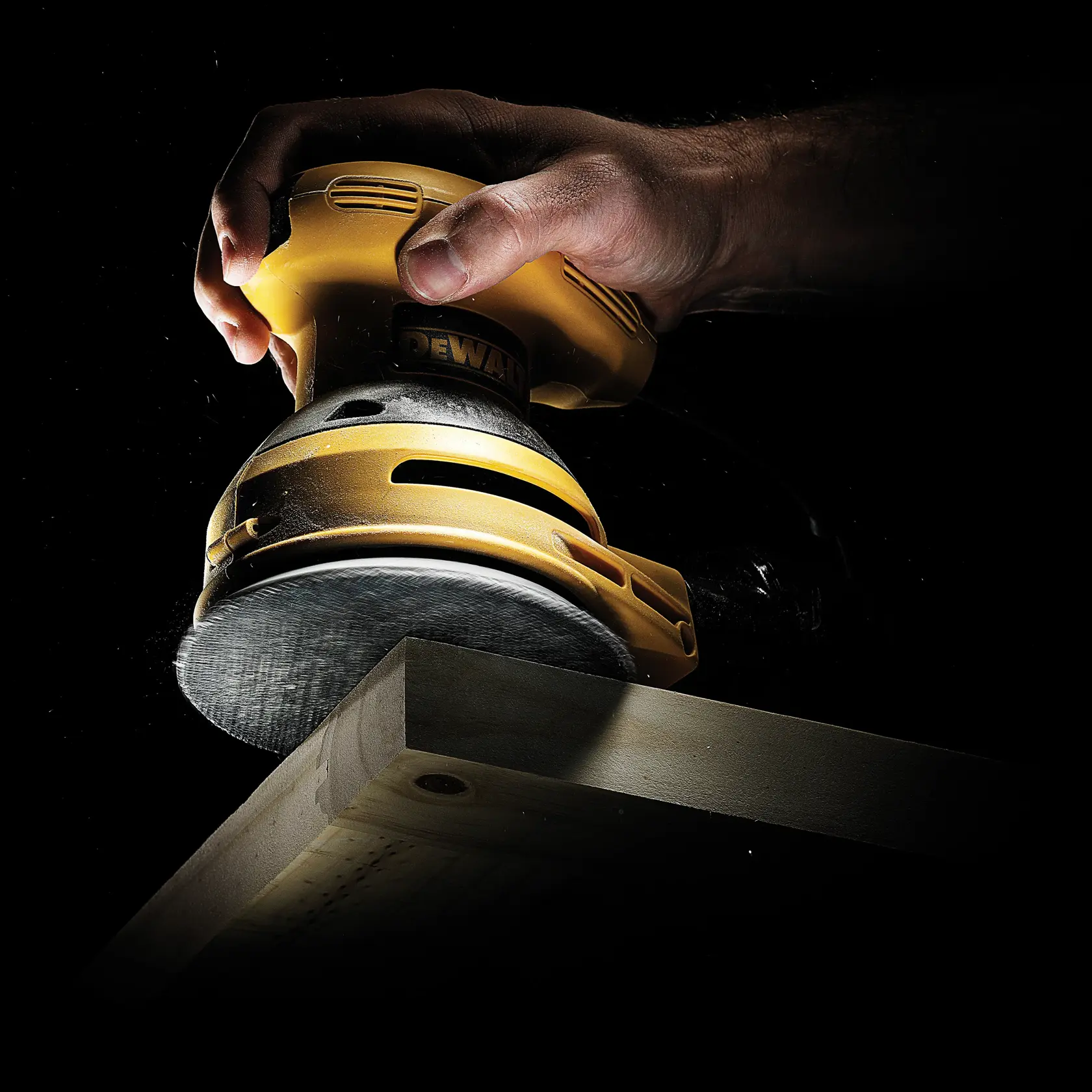 Una mano usa una lijadora orbital DEWALT para alisar el borde de una tabla de madera sobre fondo oscuro.