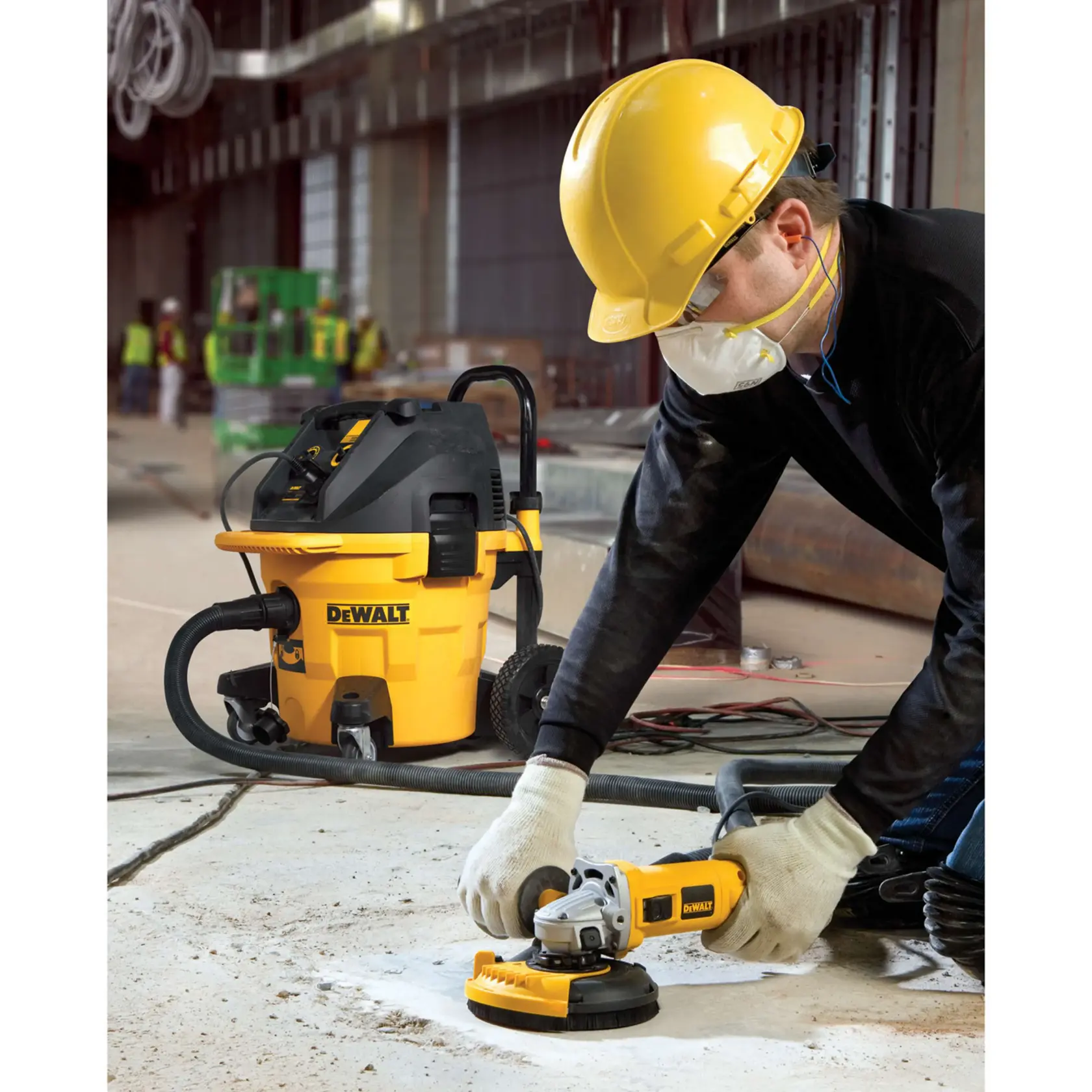 Un trabajador de la construcción con casco amarillo, guantes y mascarilla utiliza una herramienta de esmerilado DEWALT DWE46150 sobre un suelo de concreto, mientras al fondo se ve una aspiradora para polvo DEWALT amarilla. La escena ocurre en un entorno industrial o de construcción.