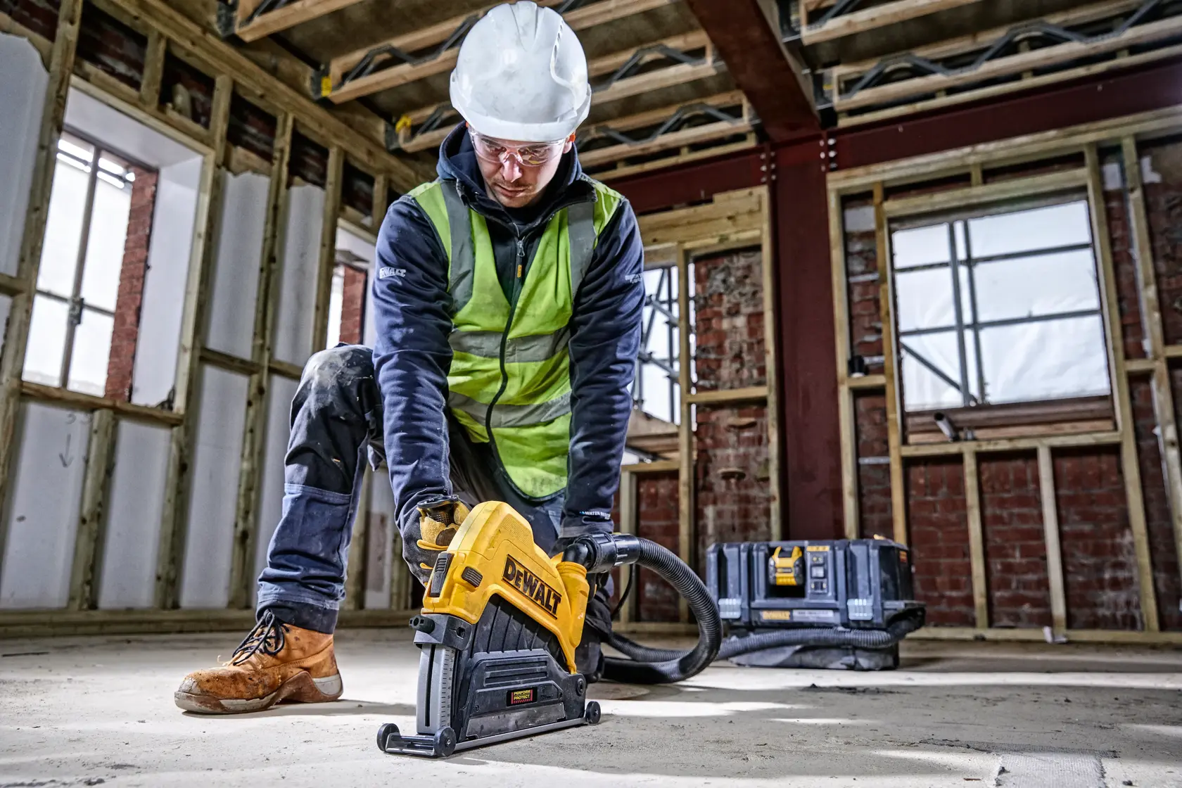 Trabajador usa herramienta DEWALT en el suelo de obra, con equipo de seguridad. Caja de herramientas atrás.