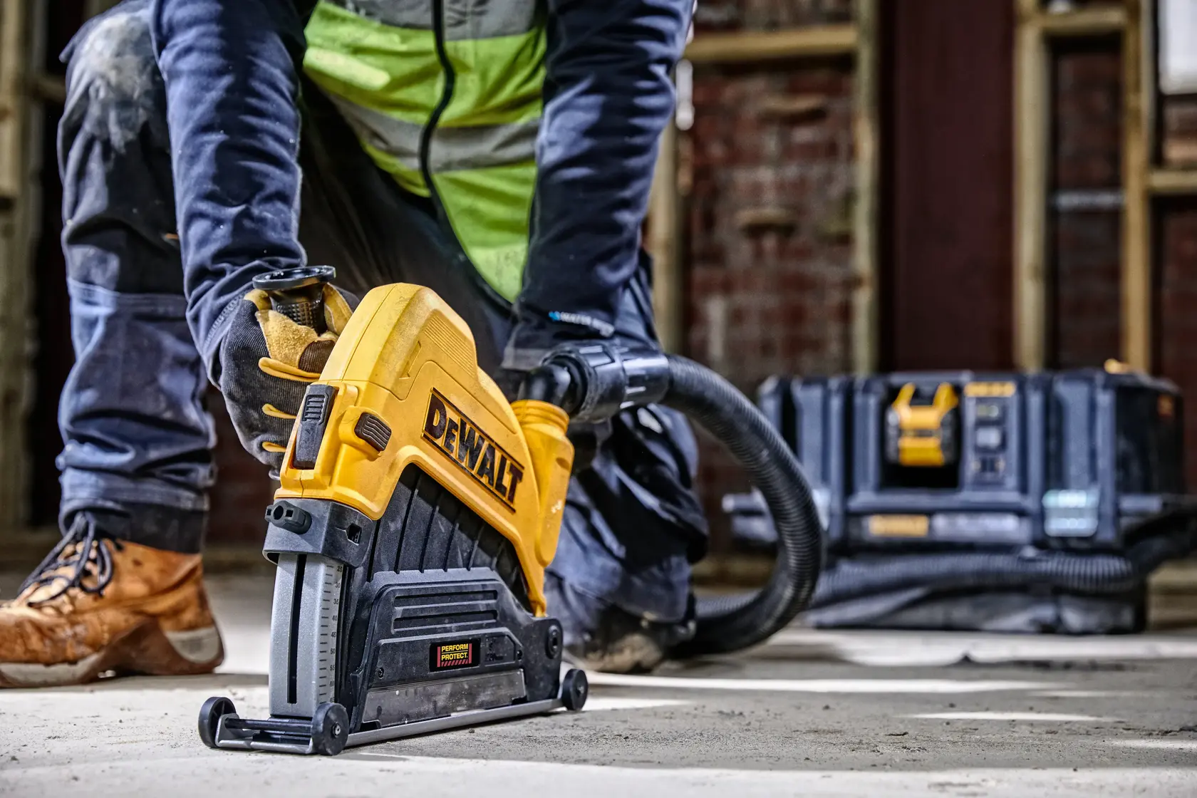 Trabajador usa herramienta DEWALT en el suelo; caja y manguera visibles en el fondo.