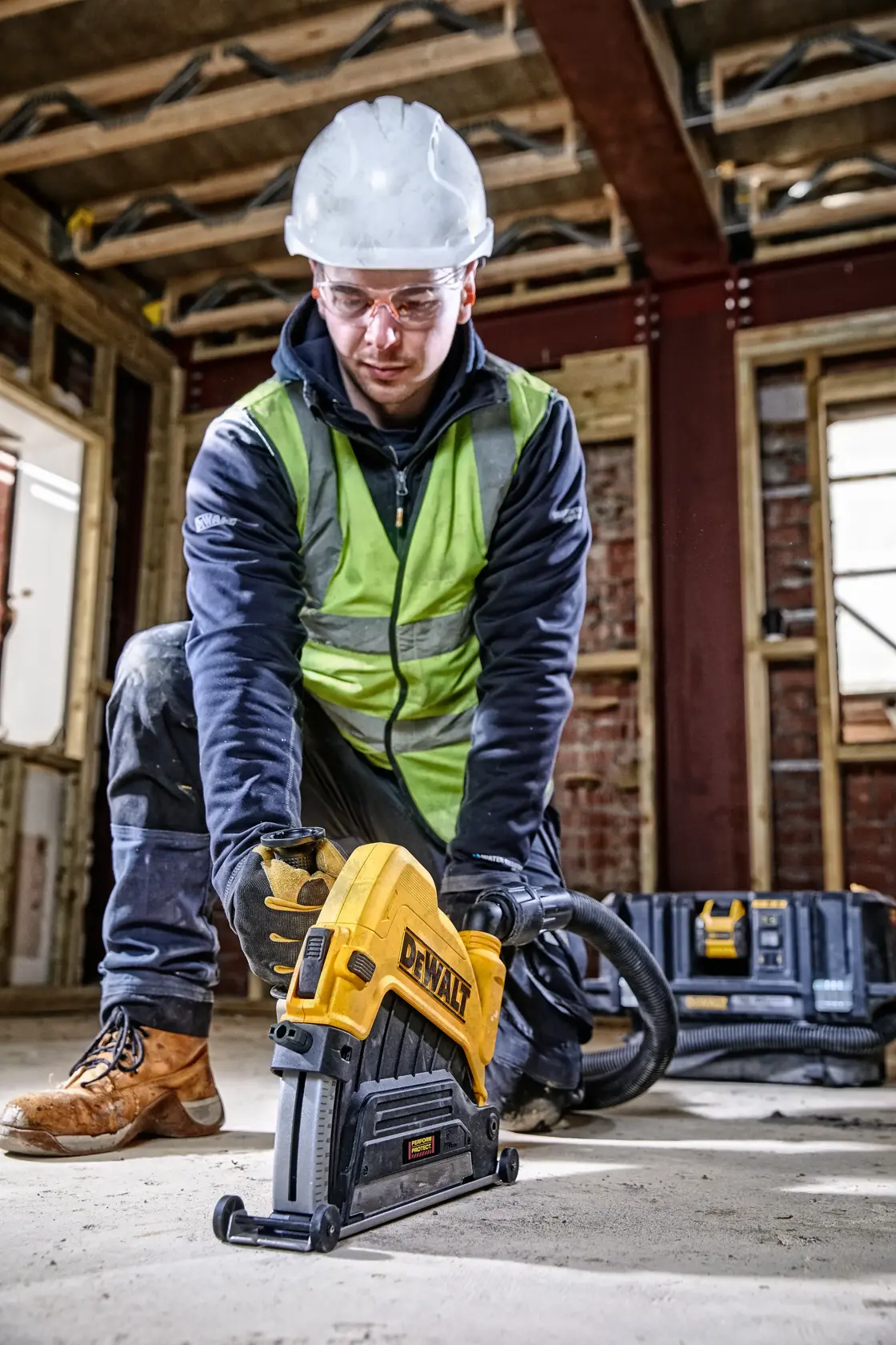 Trabajador usando herramienta DEWALT en el suelo del sitio, con equipo de seguridad y guantes.