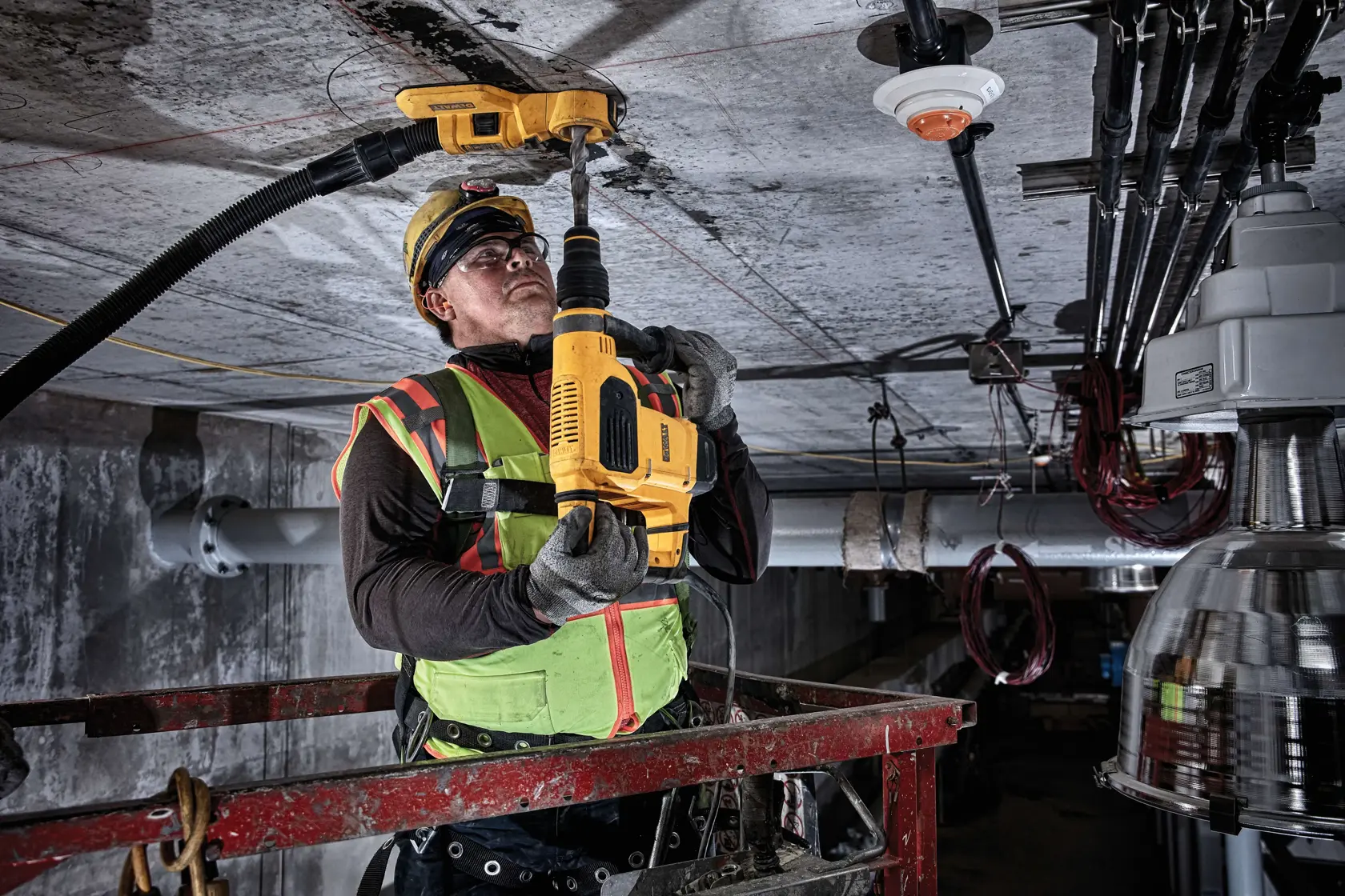 Un trabajador de la construcción con equipo de seguridad utiliza una herramienta DEWALT DWH050 para perforar el techo en un entorno industrial. El trabajador está de pie sobre una plataforma elevadora y un accesorio de extracción de polvo está conectado a la herramienta.