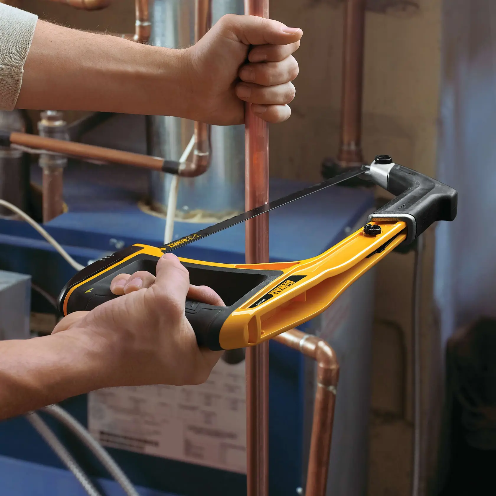 Sierra manual DEWALT cortando tubo de cobre, sostenida por dos manos en taller con tuberías visibles.