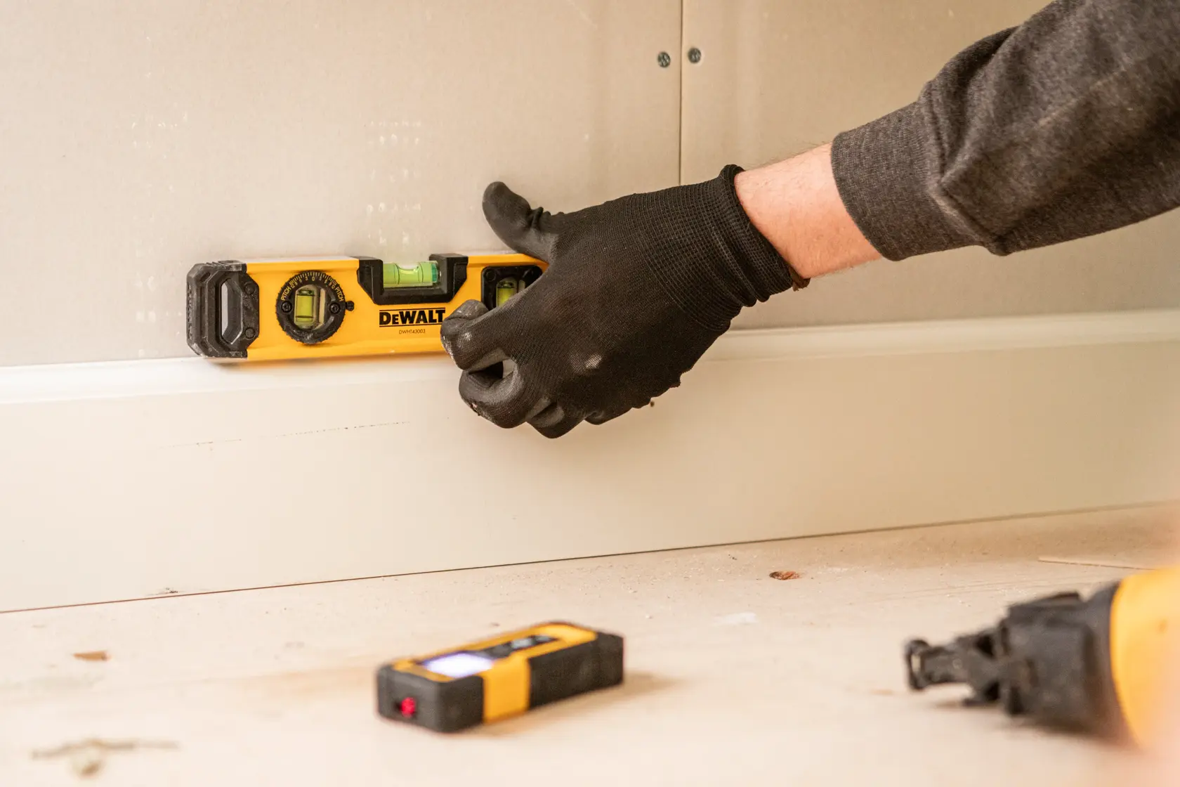 Persona usando nivel DEWALT en el zócalo de la pared con guantes negros. Hay más herramientas en el suelo.