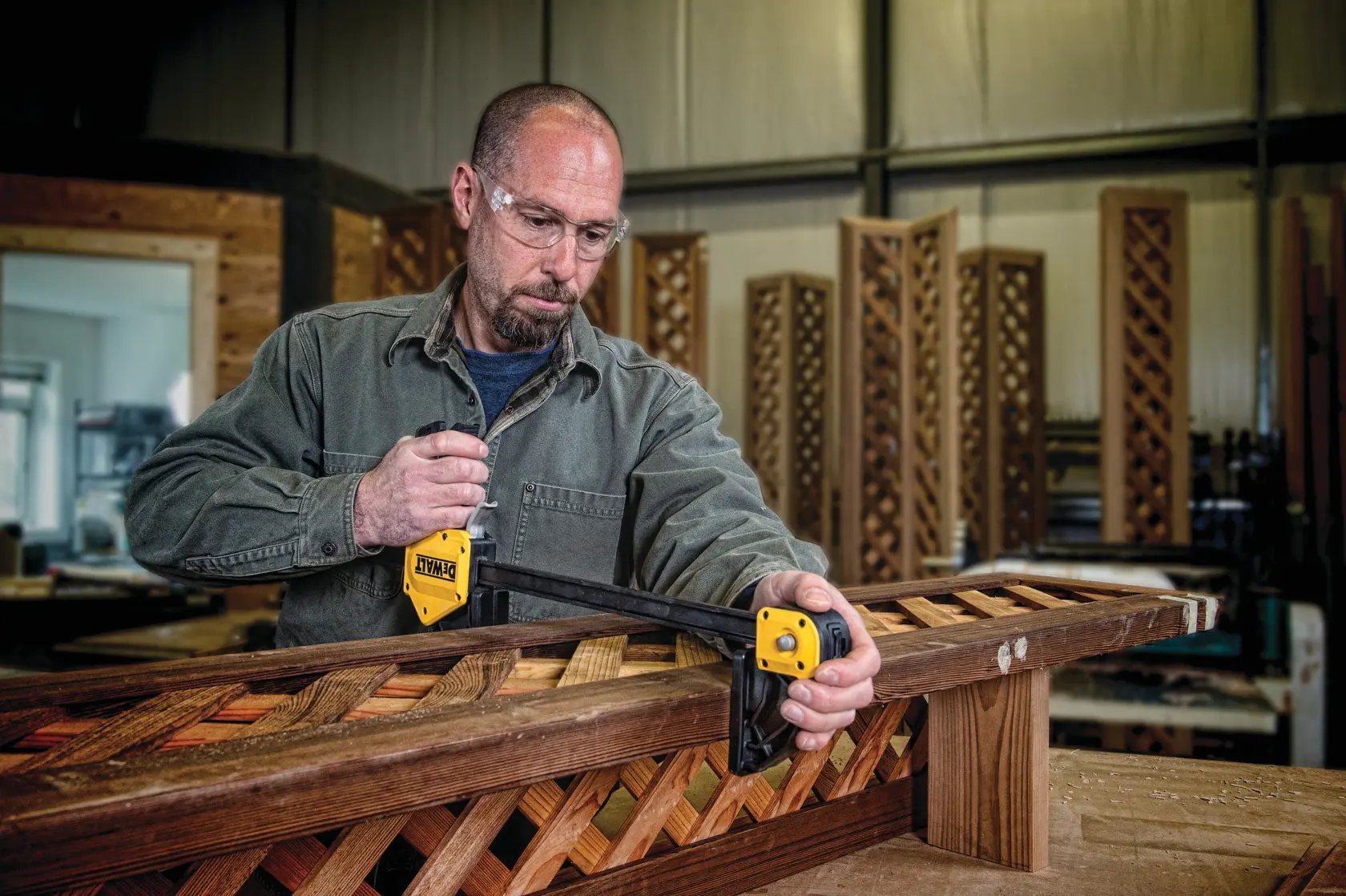 Persona usa una abrazadera DEWALT para fijar un marco de madera en taller. Rostro desenfocado.