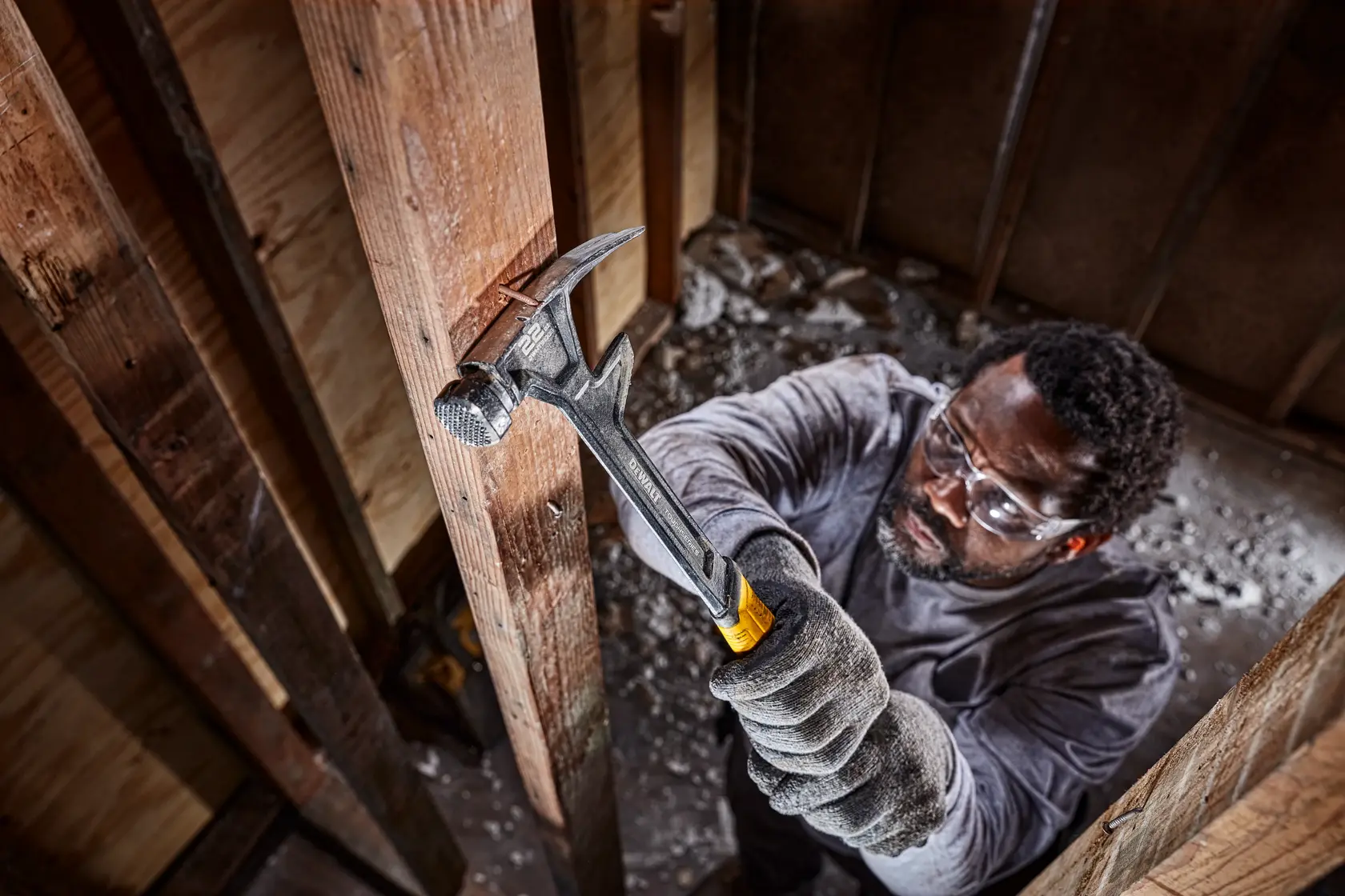 Eine Person mit Handschuhen benutzt einen DEWALT Hammer mit gelb-schwarzem Griff, um einen Holzbalken in einem Gebäude im Bau zu schlagen. Das Gesicht der Person ist unscharf.