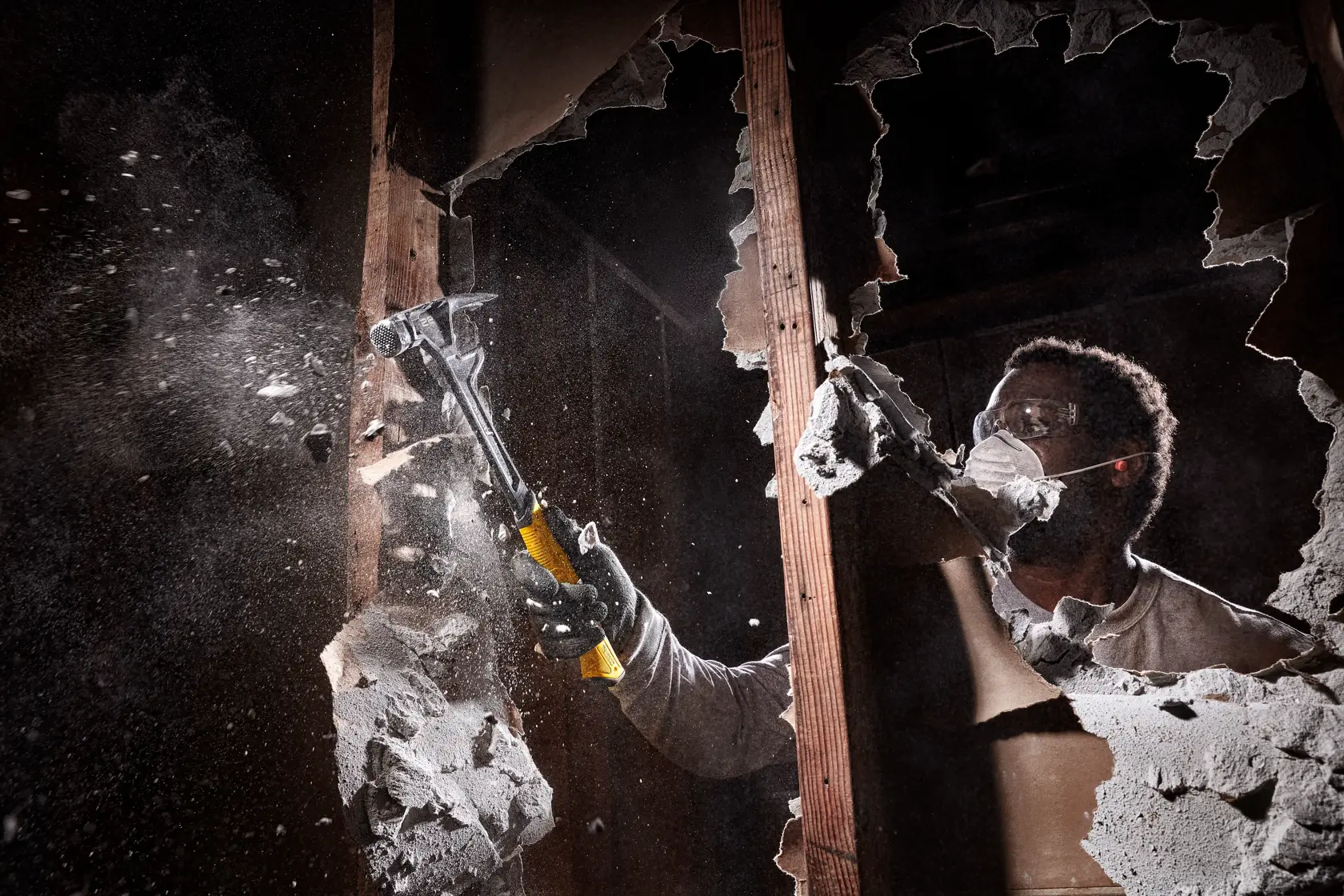 Eine Person mit Handschuhen verwendet einen DEWALT Hammer, um eine Trockenbauwand zu zerbrechen, wobei Staub und Trümmer aufwirbeln.