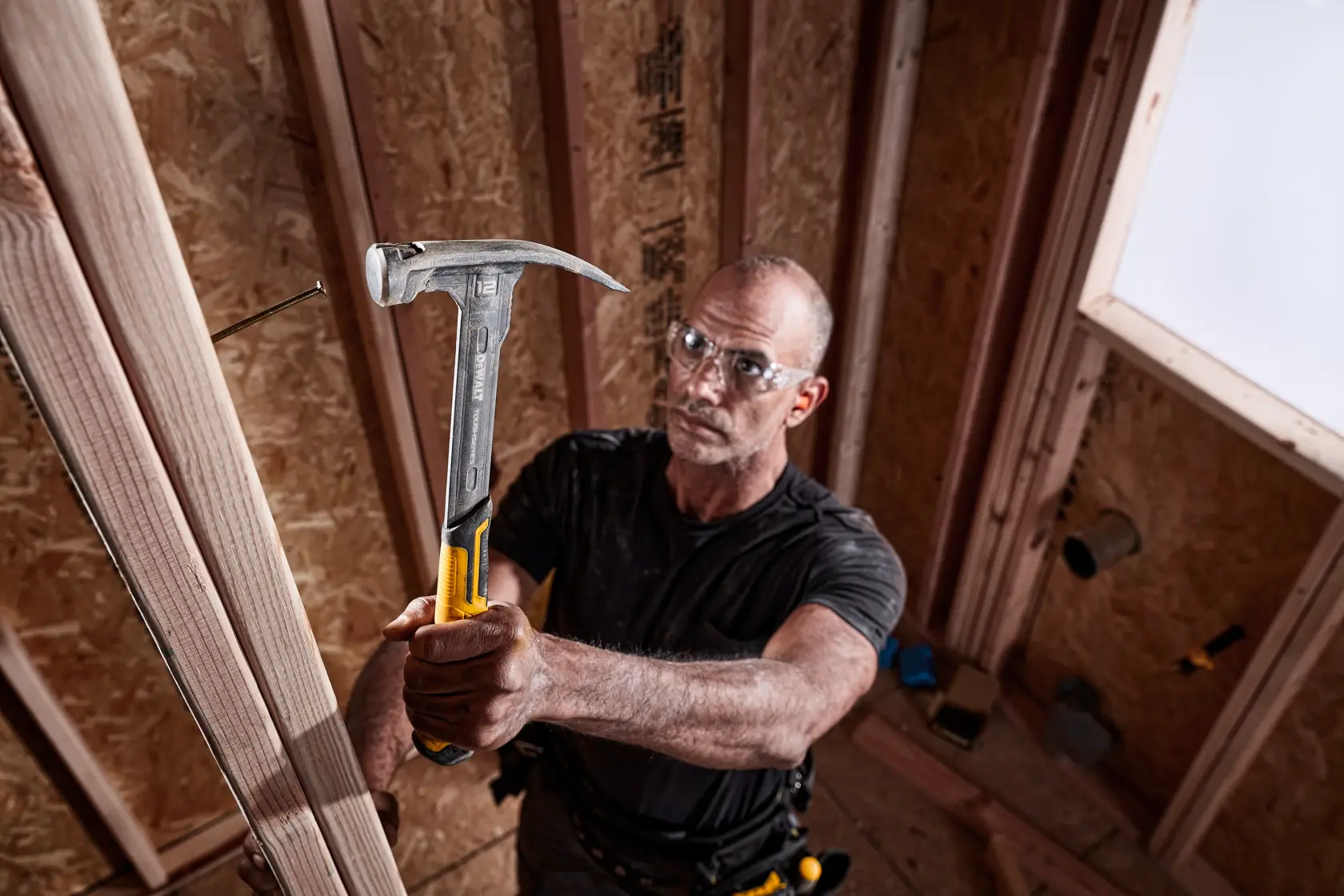 Eine Person verwendet einen DEWALT Hammer (Modell DWHT51135-0), um einen Nagel in einen Holzrahmen in einem teilweise konstruierten Raum zu schlagen. Der Hammer hat einen gelben und metallischen Griff.