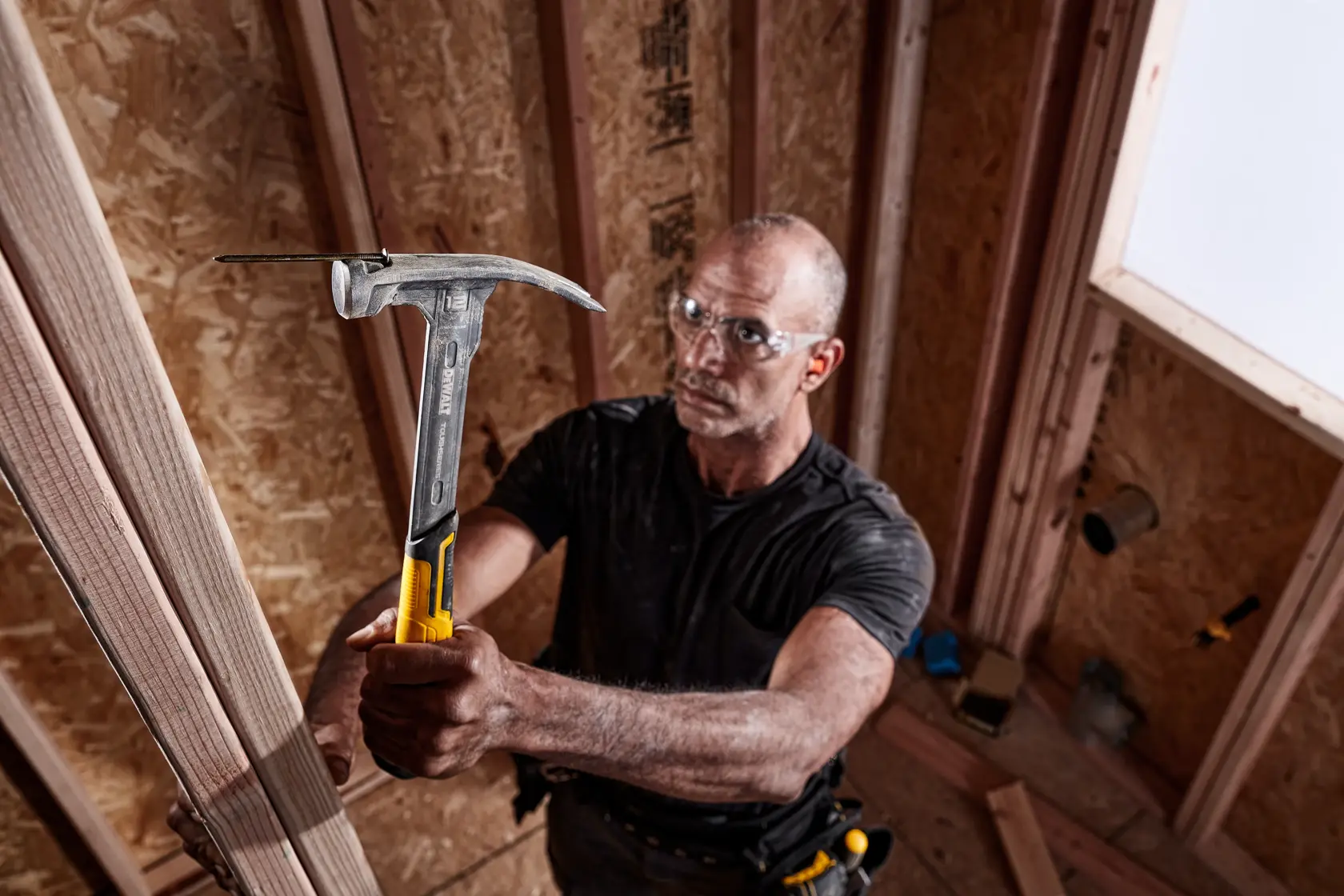 Eine Person verwendet einen DEWALT Hammer (DWHT51135-0), um an einem Holzrahmen auf einer Baustelle zu arbeiten. Der Hammer hat einen gelben und schwarzen Griff und die Person trägt einen Werkzeuggürtel.
