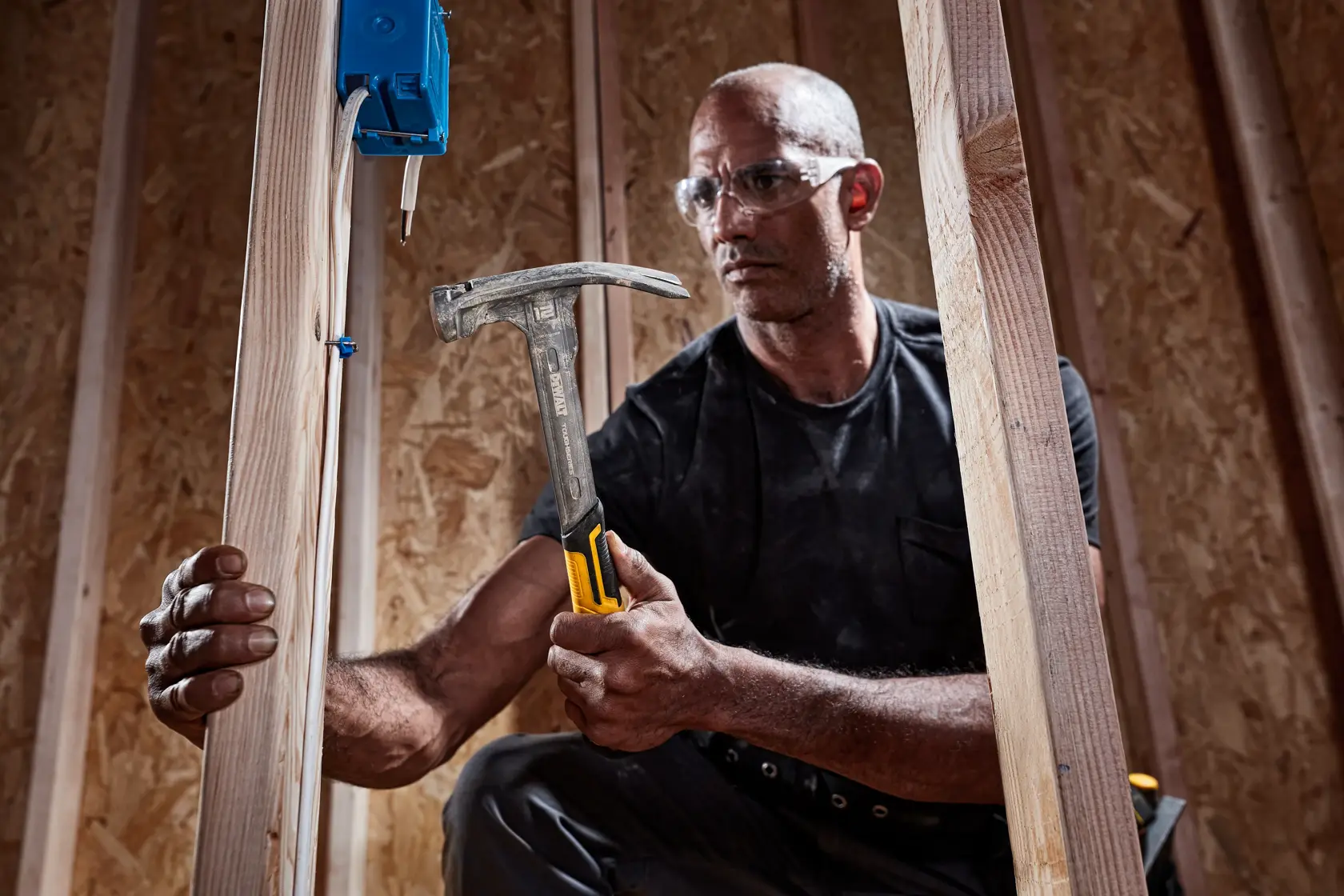 Eine Person hält einen DEWALT-Klauenhammer mit gelb-schwarzem Griff und arbeitet an einem Holzrahmenbau. Das Gesicht ist unscharf und die Person scheint eine elektrische Box an Holzbalken anzubringen.
