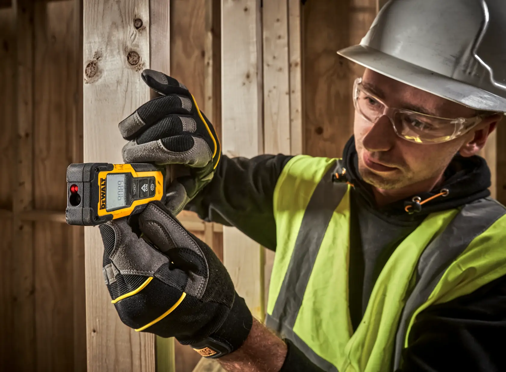 Trabajador usa DEWALT DWHT77100 medidor láser en marco de madera, con guantes y equipo de seguridad.