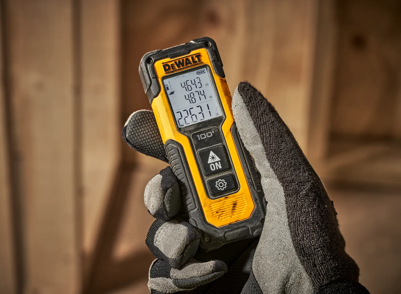 Mano enguantada sostiene medidor láser DEWALT con pantalla digital en espacio de trabajo de madera.