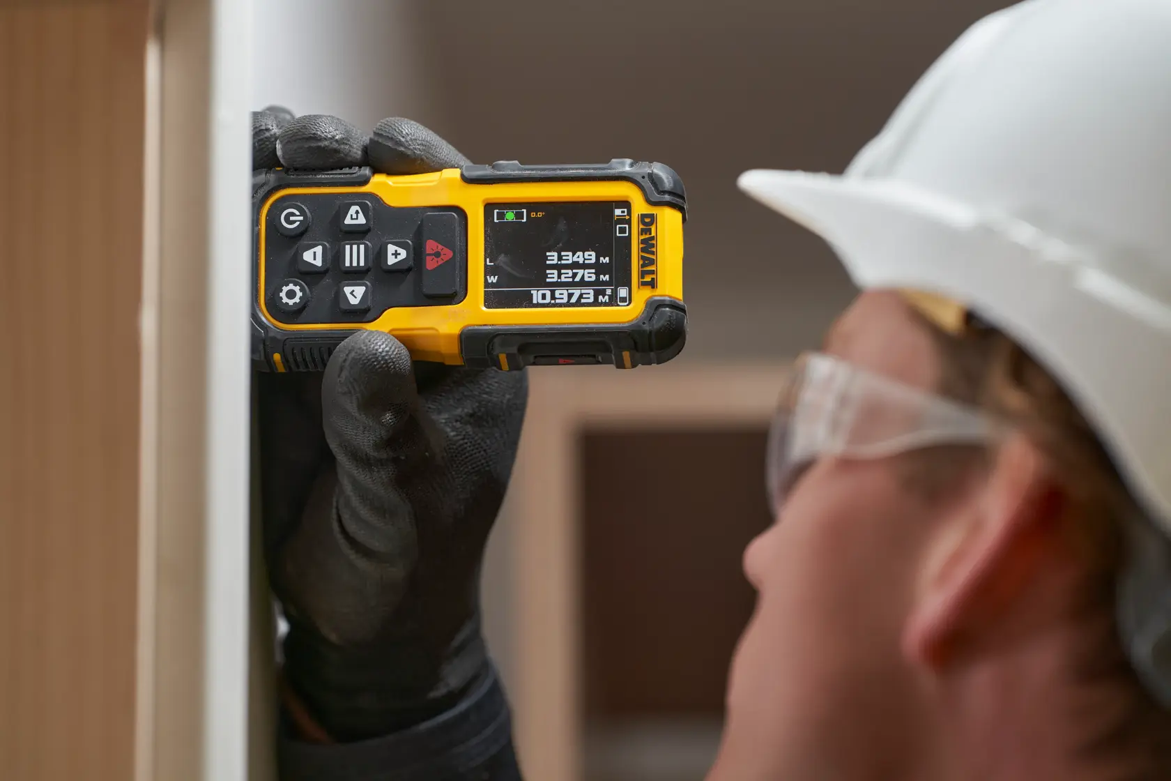 Gloved hand holds DEWALT laser distance measurer against wall; worker wears a white safety helmet.