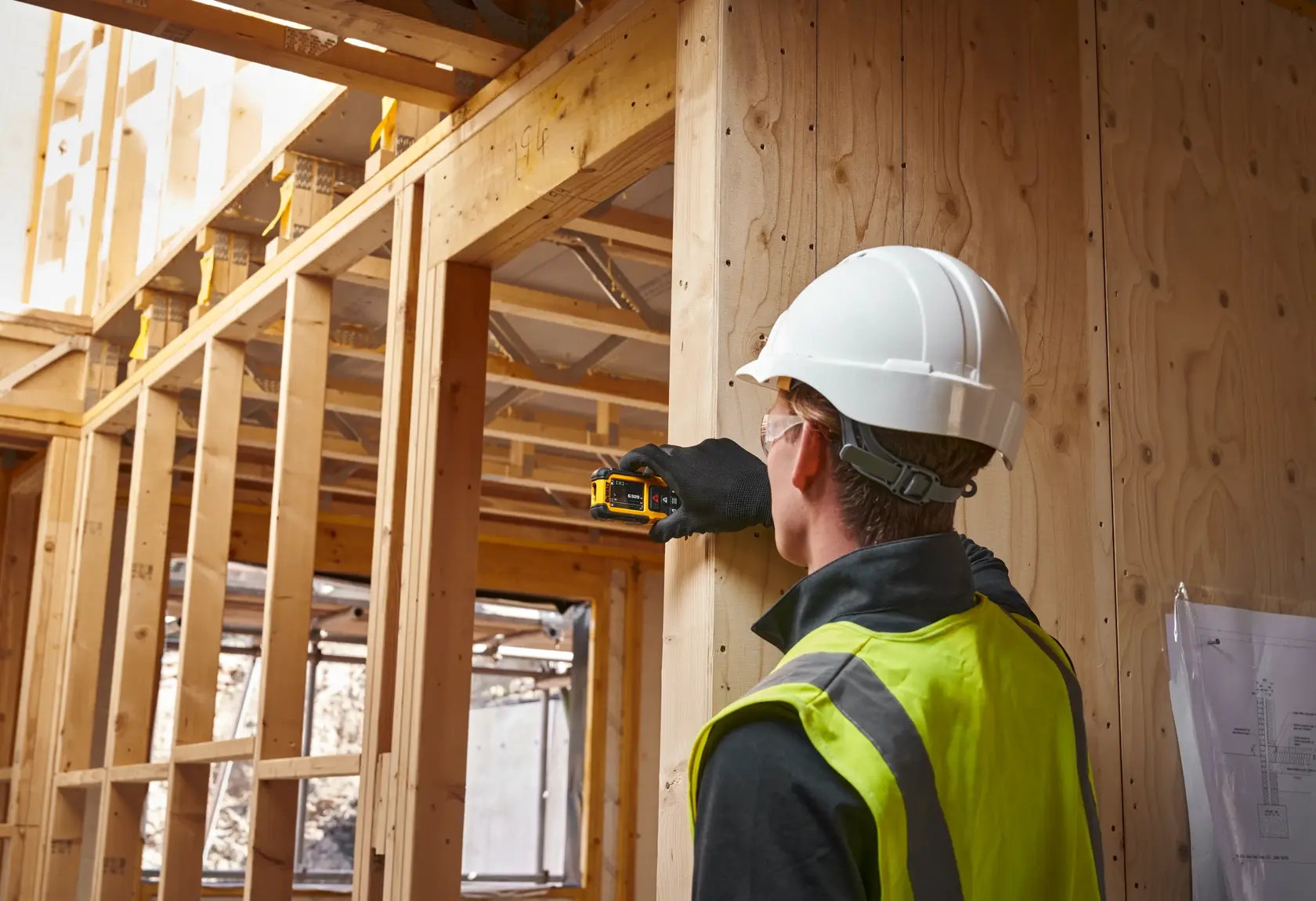 Worker uses DEWALT laser measurer on wood frame at construction site, wearing safety gear and reflective vest.