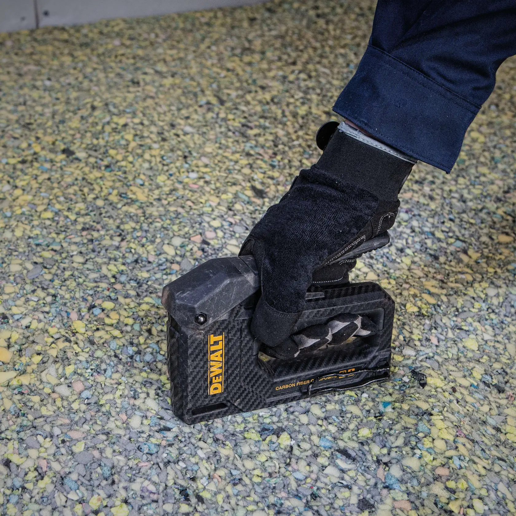 Eine behandschuhte Hand verwendet einen DEWALT Kohlefaser-Tacker, um Material auf einer gesprenkelten Oberfläche zu befestigen.