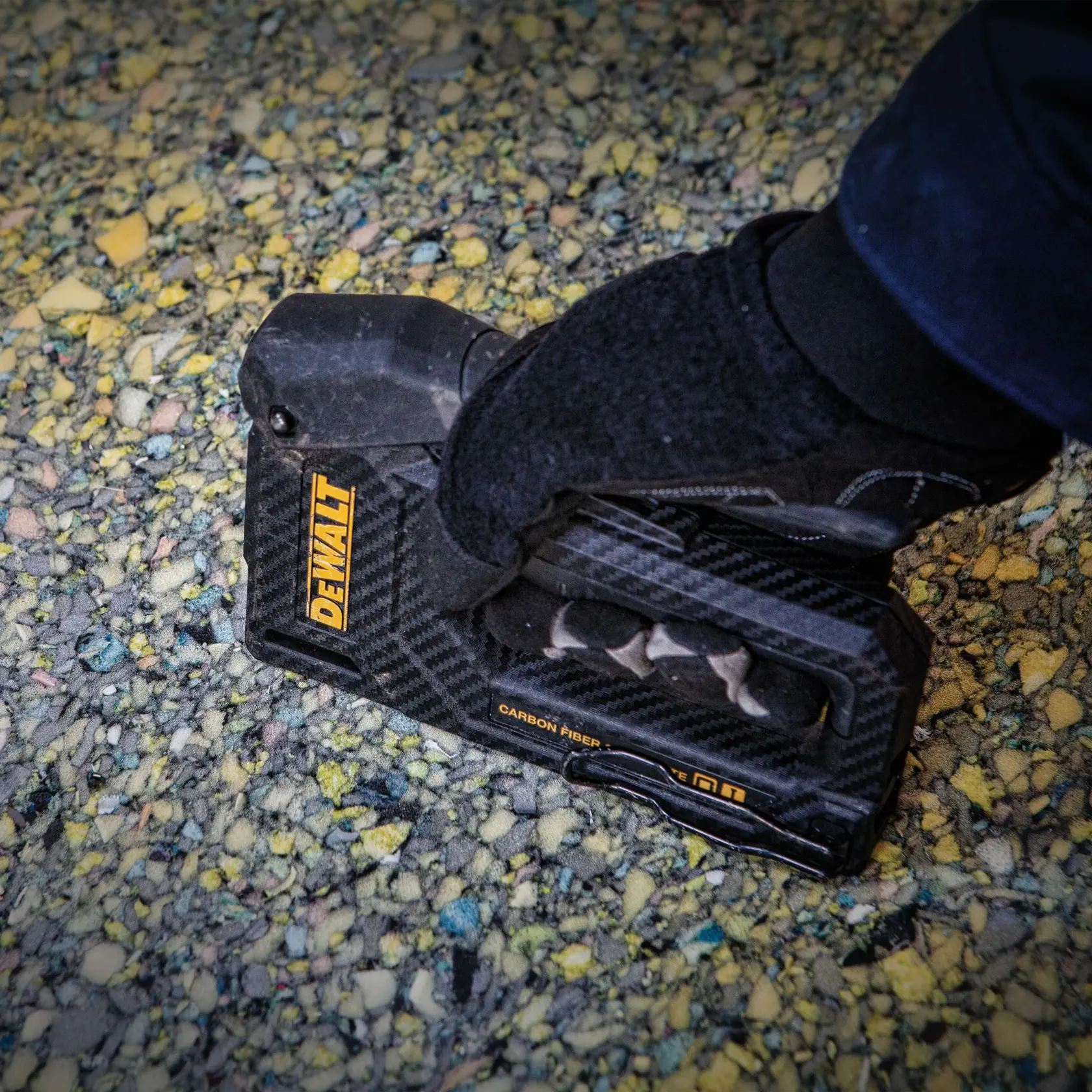 Nahaufnahme einer behandschuhten Hand, die ein DEWALT-Handwerkzeug aus Karbonfaserverbundstoff auf einer texturierten Oberfläche mit kleinen, bunten Steinen hält.