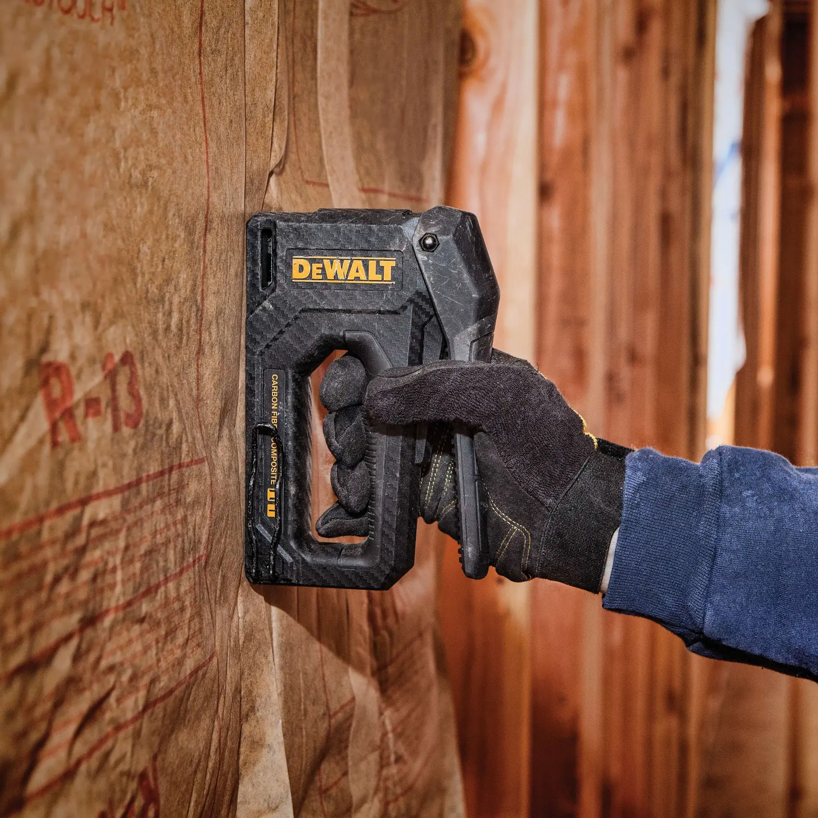 Eine Person mit einem schwarzen Arbeitshandschuh verwendet einen DEWALT-Handtacker, um Isoliermaterial an einer Holzwand zu befestigen.