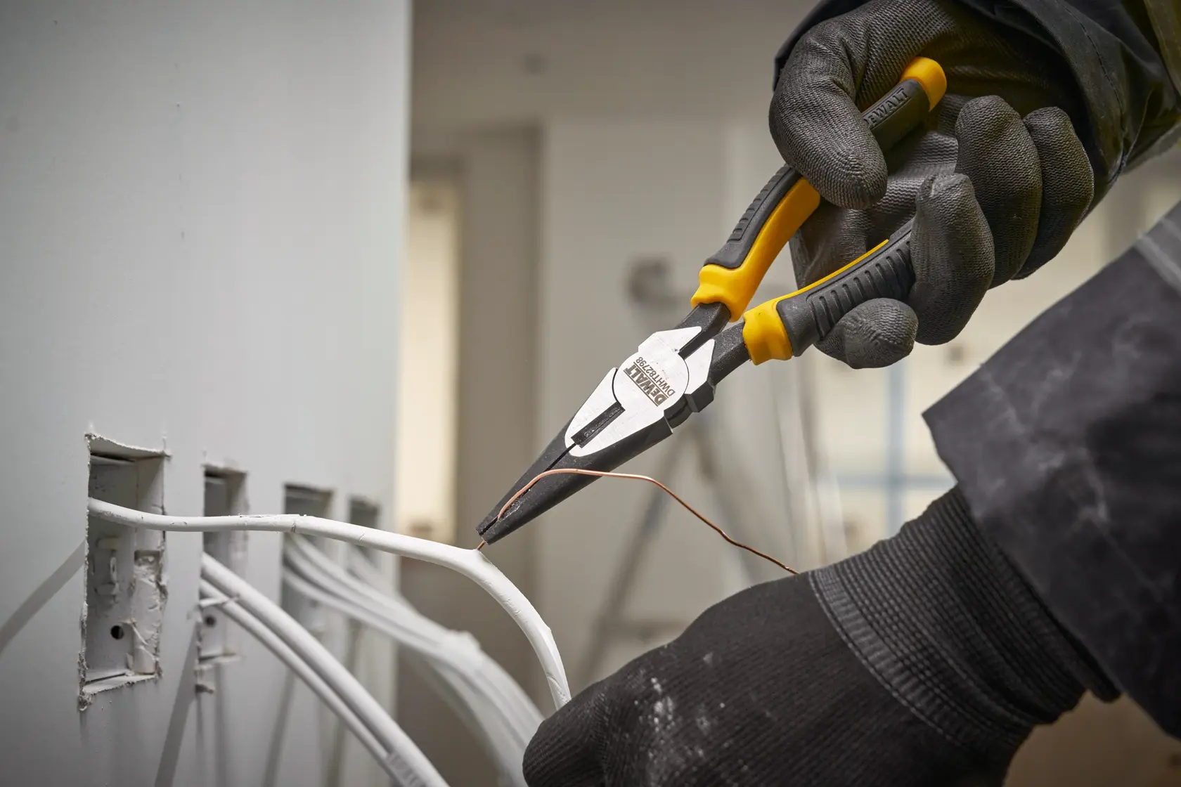 Gloved hand uses DEWALT pliers to handle wire near electrical wall outlets during installation.