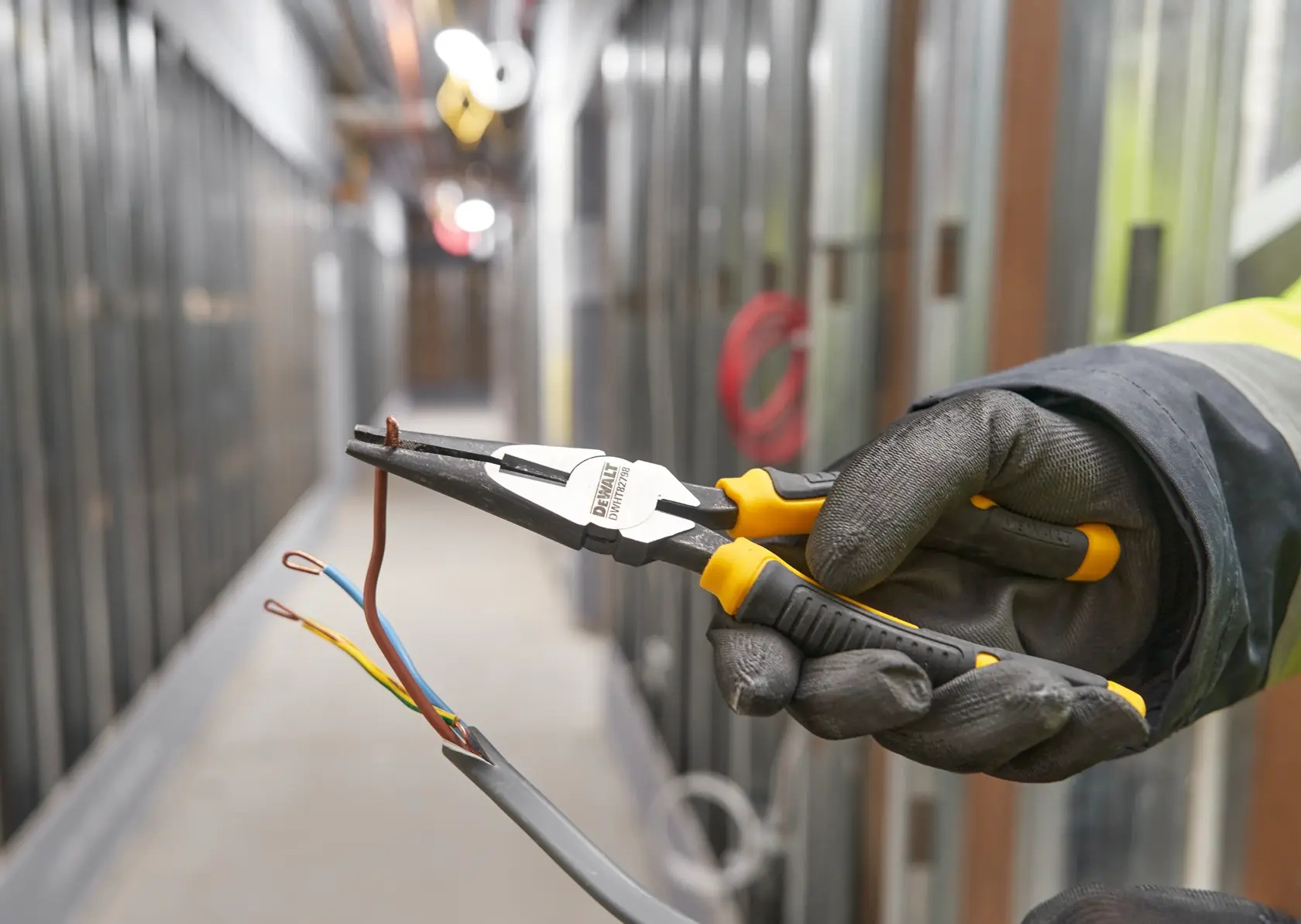 Gloved hand using DEWALT pliers to grip electrical wires in an industrial hallway.