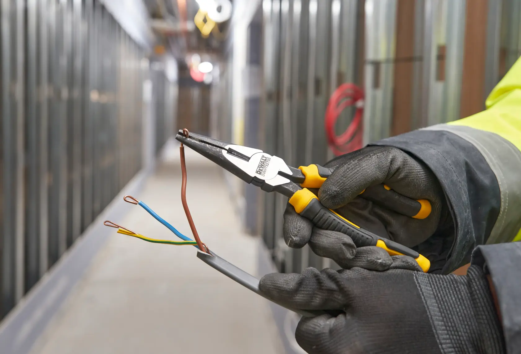 Gloved hands use DEWALT pliers to bend electrical wires in a hallway under construction.