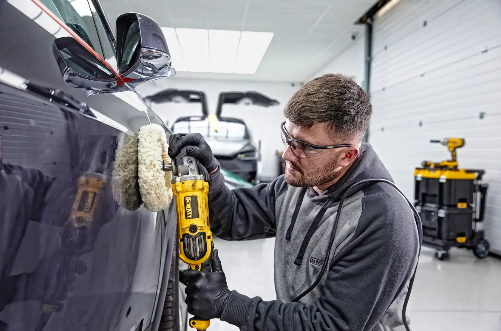 A person is polishing the hood of a red sports car with a DEWALT DWP849X polisher in a garage.