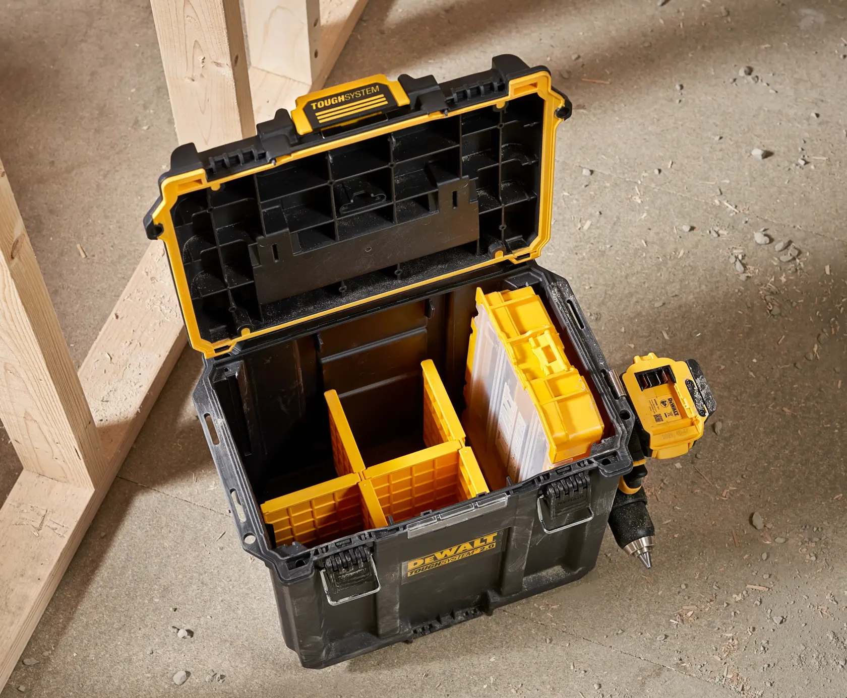 Caja de almacenamiento DEWALT ToughSystem abierta con compartimentos amarillos y un taladro adjunto en el lateral, situada sobre un suelo de hormigón junto a un marco de madera.