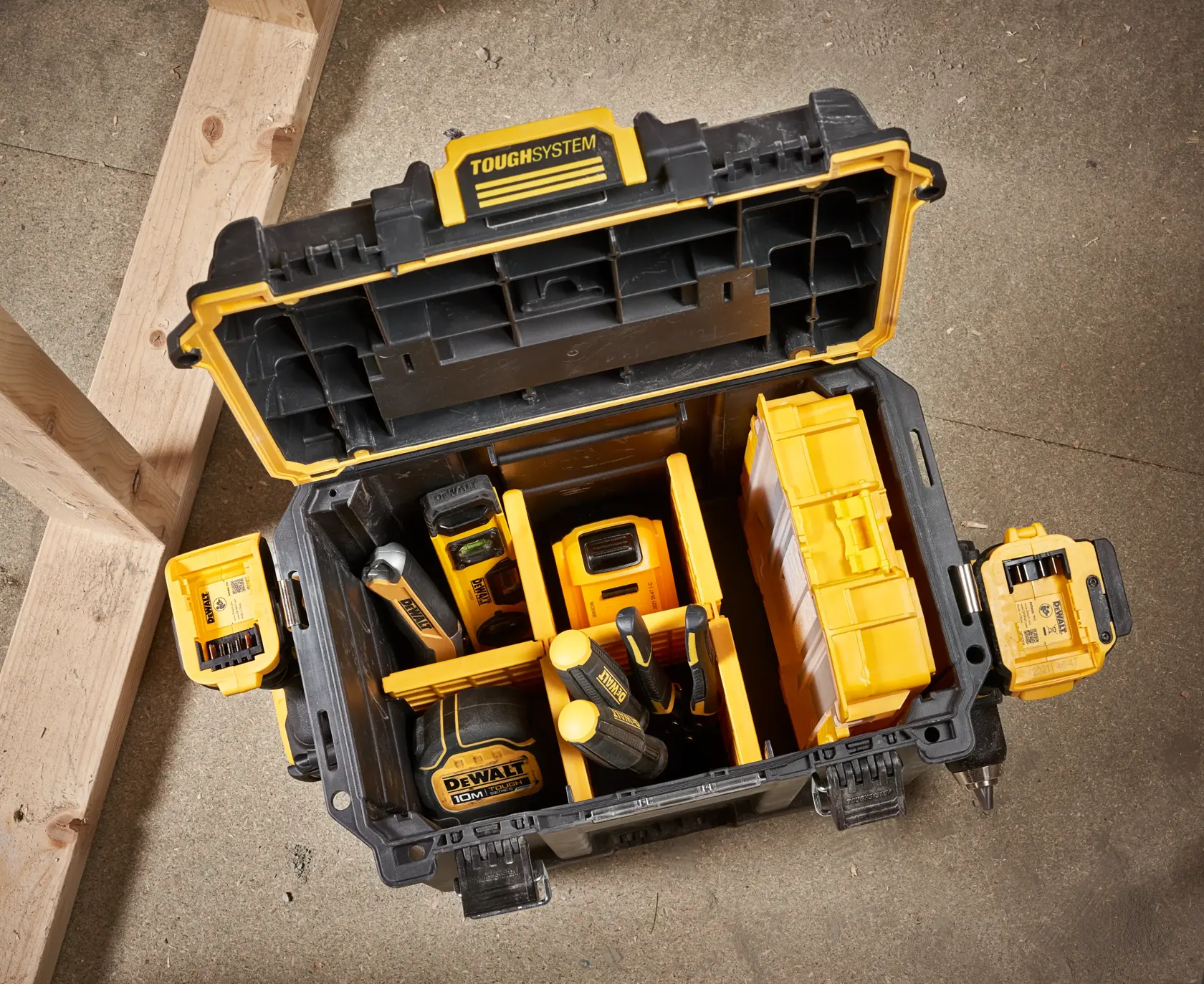 Caja de herramientas DEWALT ToughSystem abierta que contiene varias herramientas y accesorios amarillos y negros, colocada sobre un suelo de cemento junto a vigas de madera.