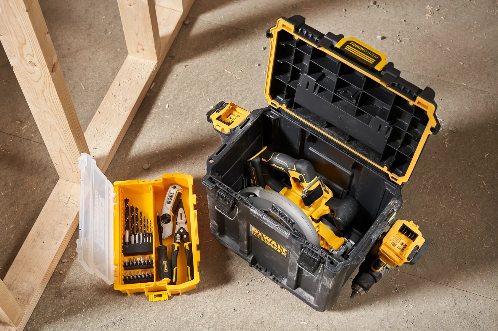 Una caja de almacenamiento de herramientas DEWALT con la tapa abierta, que contiene una sierra circular. Al lado, un organizador pequeño amarillo y transparente con brocas y alicates, ambos sobre el suelo del taller.