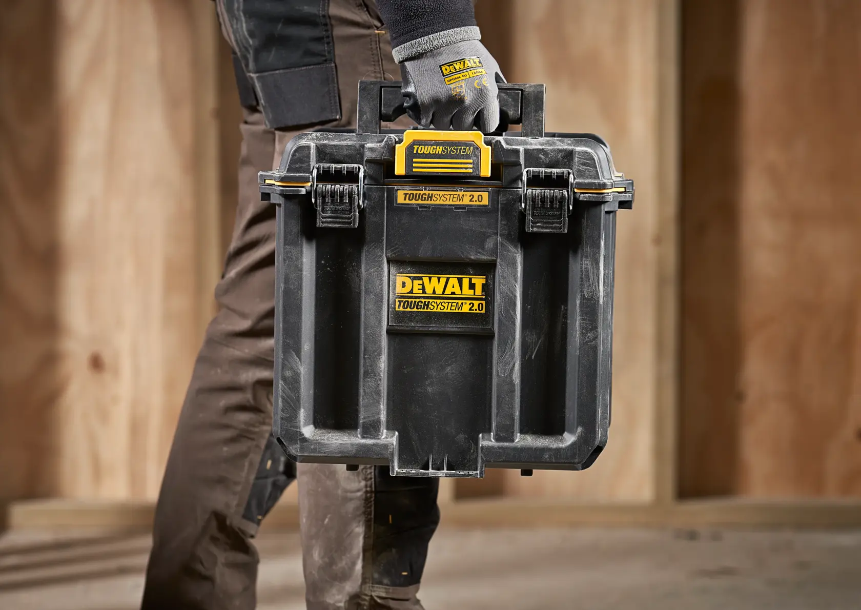 Una persona con guantes de trabajo transporta una caja de almacenamiento negra DEWALT TOUGHSYSTEM 2.0 con etiquetas amarillas, diseñada para organizar y transportar herramientas, en un entorno de construcción con paredes de madera.