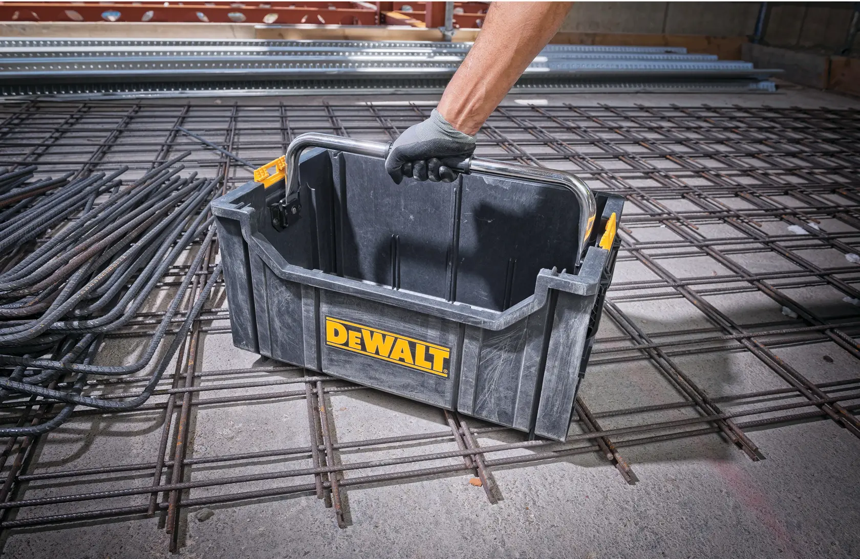 Person lifts a DEWALT plastic tool box with a handle, placed on a metal grid in a construction site.