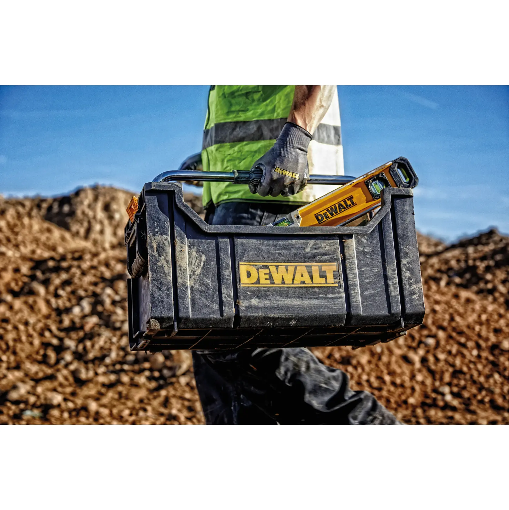 Worker carrying a DEWALT toolbox with tools outdoors at a construction site.