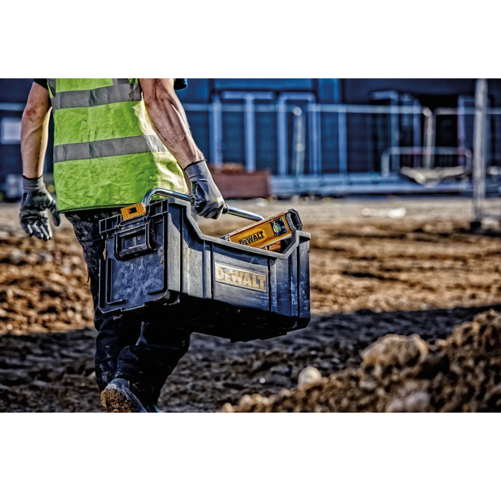 Person carries a DEWALT toolbox with tools on a construction site, wearing gloves and a reflective vest.