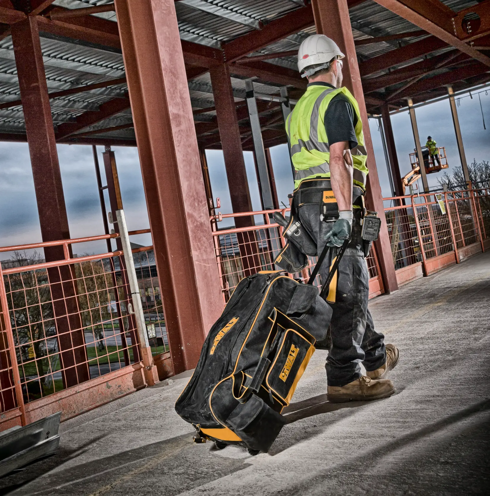 Trabajador arrastra bolsa DEWALT con ruedas en obra, vigas de acero y equipo de seguridad visibles.