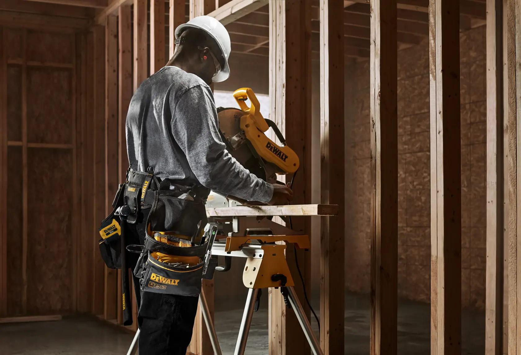 Eine Person mit Schutzhelm und Werkzeuggürtel bedient eine DEWALT Kappsäge auf einer Baustelle mit Holzrahmen.