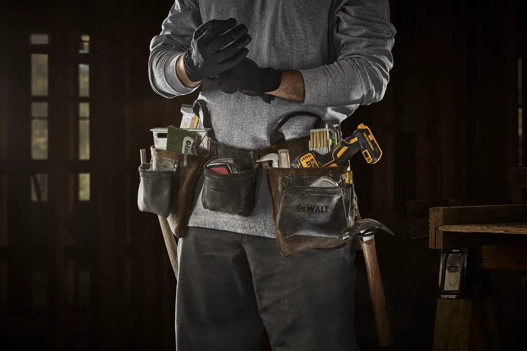 Una persona lleva un cinturón de herramientas DEWALT lleno de herramientas como cinta métrica, destornilladores, lápices y un taladro inalámbrico amarillo y negro, de pie en un taller y poniéndose guantes de trabajo negros.