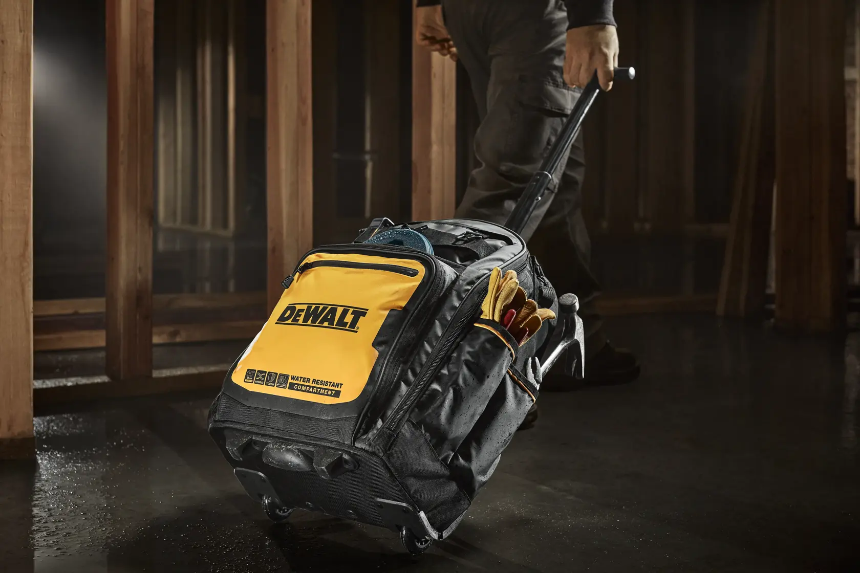 A DEWALT branded black and yellow rolling tool bag with an extendable handle, being pulled by a person in a worksite environment. The bag is equipped with exterior pockets holding work gloves and other tool accessories.