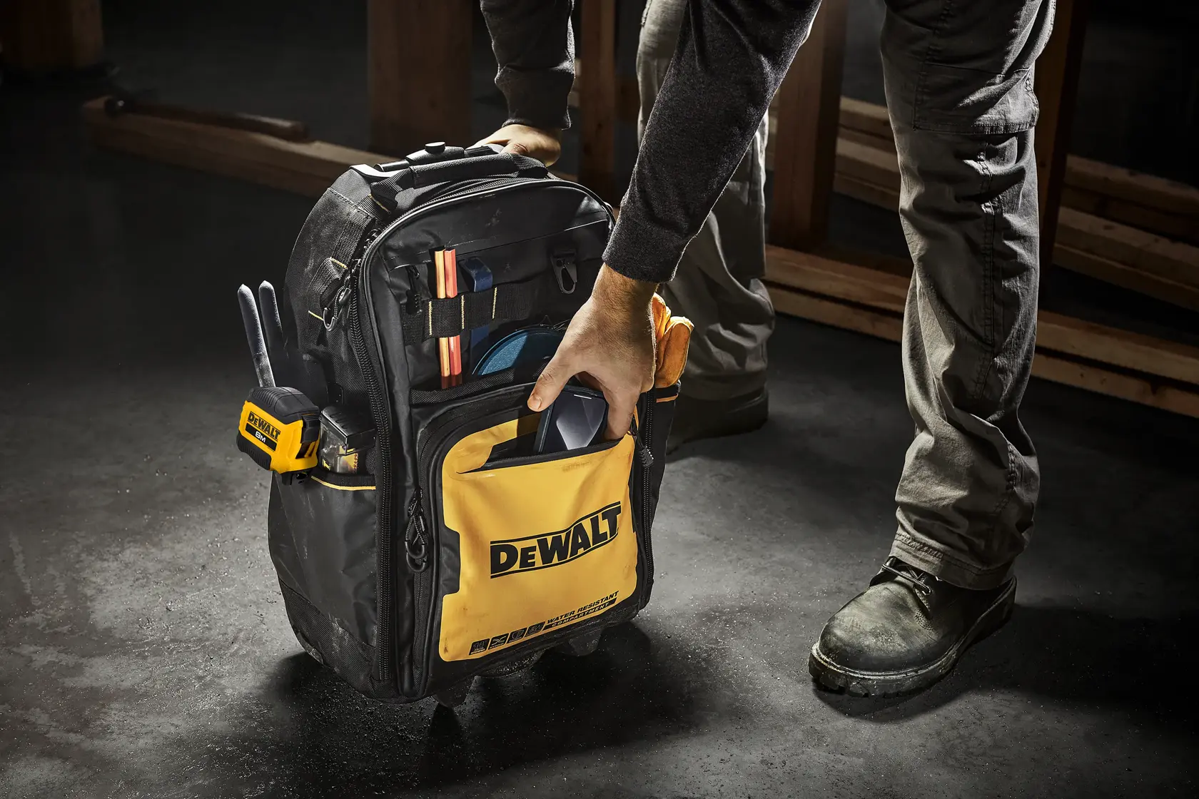 A person is holding a DEWALT branded backpack tool bag, which is filled with various tools and equipment. The bag is yellow and black, and is positioned on a workshop floor.